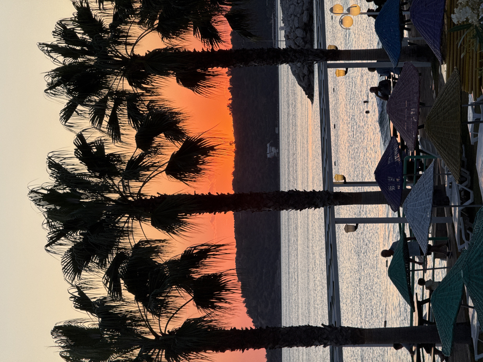 The image depicts a sunset scene at a beach resort in Menderes, Turkey. In the foreground, several palm trees are silhouetted against a vibrant orange and pink sky. Below the palm trees, beach umbrellas and lounge chairs are arranged on a wooden deck, with some people seated at tables and others walking around. The sea is calm, with gentle waves lapping at the shore. In the background, a tree-covered hill rises from the water, with a few buildings visible on its slopes. The overall atmosphere is tranquil and relaxing, suggesting a perfect end to a day at the beach.