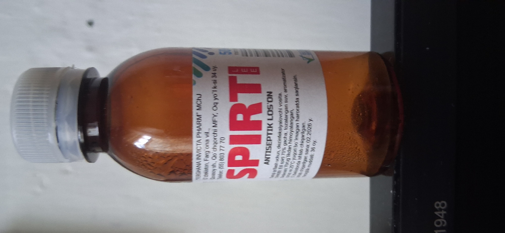 A brown glass bottle with a white cap containing a clear liquid, likely an antiseptic lotion, is photographed against a white background. The bottle has a label with text in both Latin and Cyrillic alphabets. The label indicates the product name "SPIRTI" in large red letters, along with "ANTISEPTIK LOS'ON". There is also information about the manufacturer, "TERGHANA INVICTA PHARM MCHJ", and an address in Uzbekistan: "O'zbekiston, Farg'ona vil., Quvasoy sh., Qo'qhorchi MFY". A phone number "(55) 803 77 70" is also listed. Below the product name, there is a description of its use, ingredients (including 70% spirit, distilled water, and aromatizer), storage instructions (protect from light and store at a temperature not exceeding 25°C), manufacturing date (02.2026 y.), and expiration period (36 months). The bottle is placed on a black surface with the number "1948" visible. The overall lighting suggests an indoor setting, and no specific geographical landmarks of Almalyk, Uzbekistan are identifiable in the image itself.