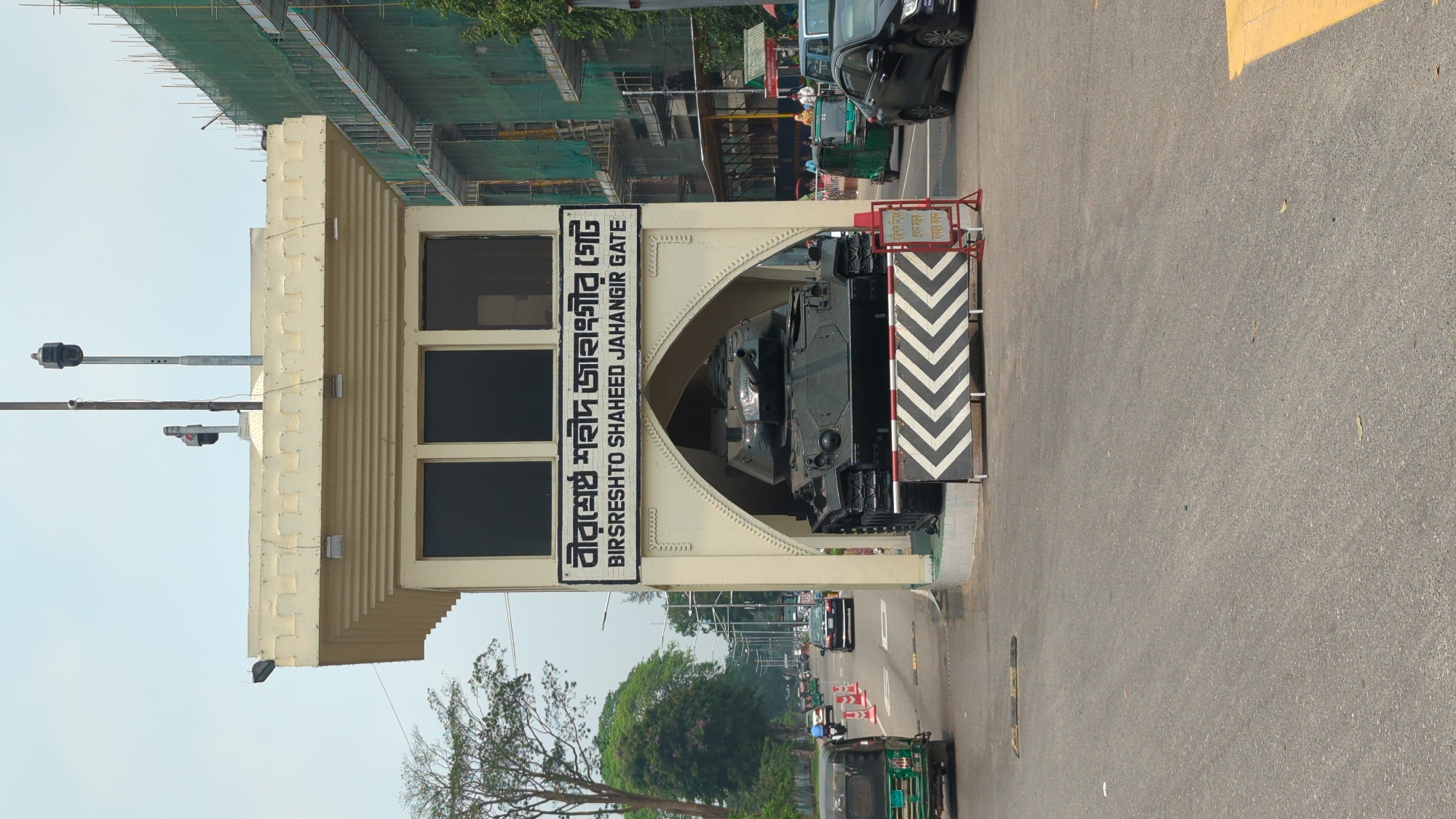 The image depicts Birshreshto Shaheed Jahangir Gate in Dhaka, Bangladesh. The gate, constructed of beige stone, straddles a road, with a black tank displayed within its arch. A sign in Bengali and English identifies the gate. A black car is visible to the right, with other vehicular traffic receding into the distance. Construction is evident on the upper left side of the frame. Traffic lights are visible on the left side of the gate. Road markings and chevron warning signs indicate traffic patterns. Trees are interspersed throughout the image.