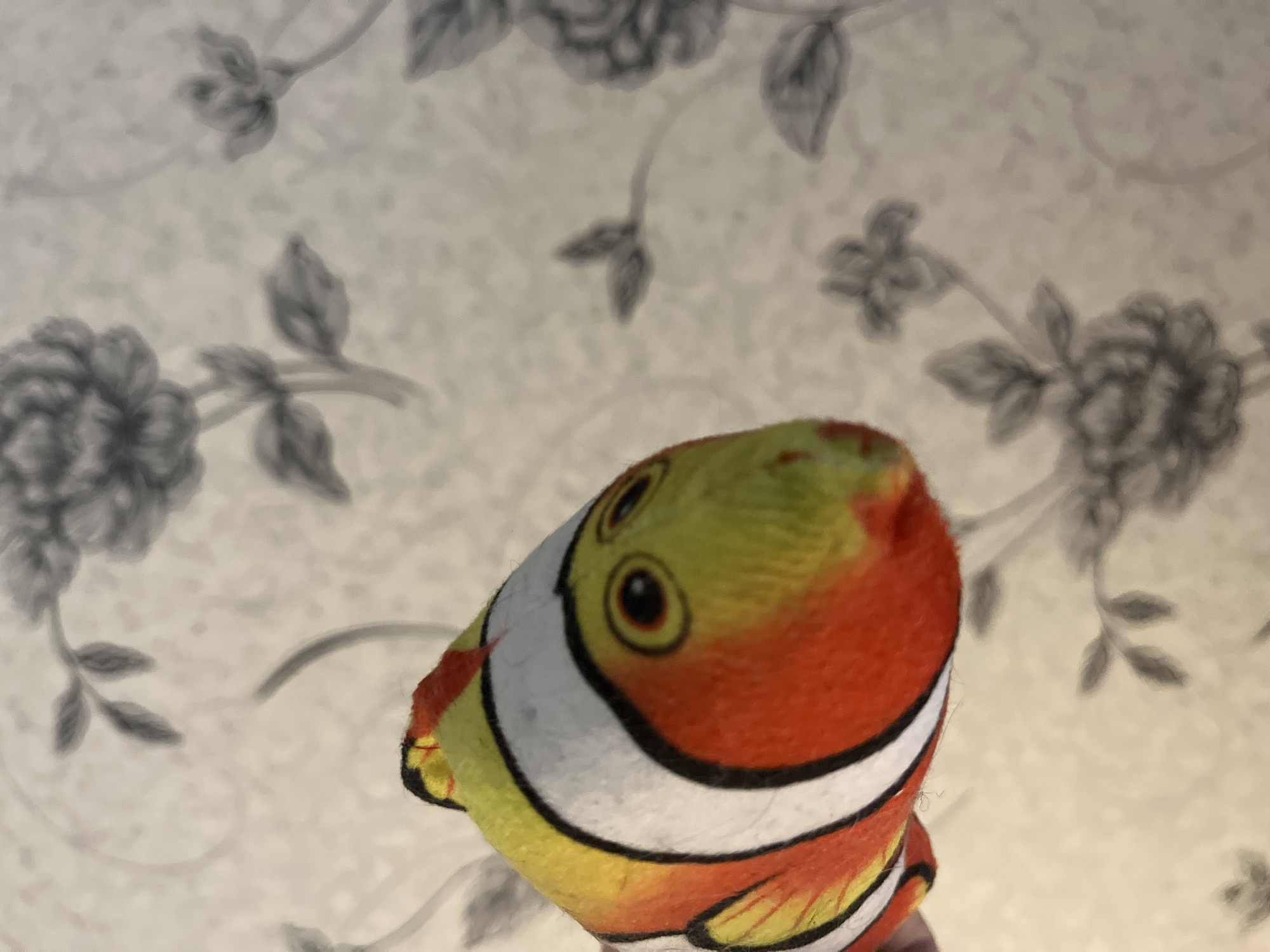 A stuffed clownfish toy is the central subject of the image. The toy is composed of orange, yellow, and white fabric and is decorated with black stripes. It features two circular eyes. The background is a wall covered with floral wallpaper featuring gray roses. The location is not definitively discernible as Chaikovsky, Russia.
