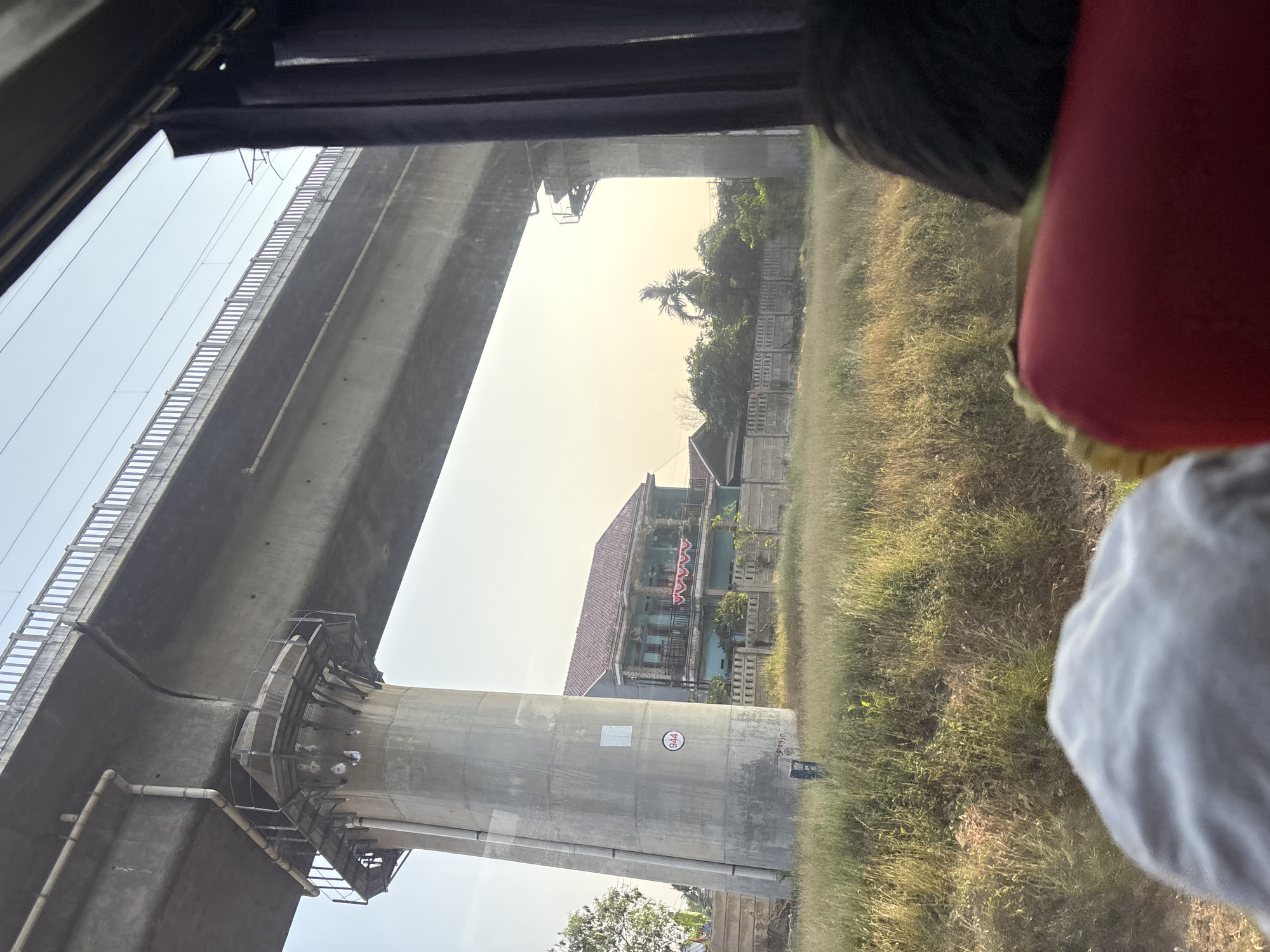 The image captures a view from a vehicle in Jayamukti, Indonesia, looking towards an elevated railway line. A massive concrete pillar supports the overpass, with a metal platform and railings visible on its upper section. Below the overpass, a two-story house with a red-tiled roof is situated behind a concrete fence. The house has blue walls and a balcony adorned with red and white bunting. The foreground is dominated by dry, tall grass. In the bottom right corner, the heads of two passengers are visible, one with dark hair and the other with a red headrest in front. The sky is clear with a hint of haze, and the overall lighting suggests it is late afternoon. The environment appears to be semi-urban or rural.