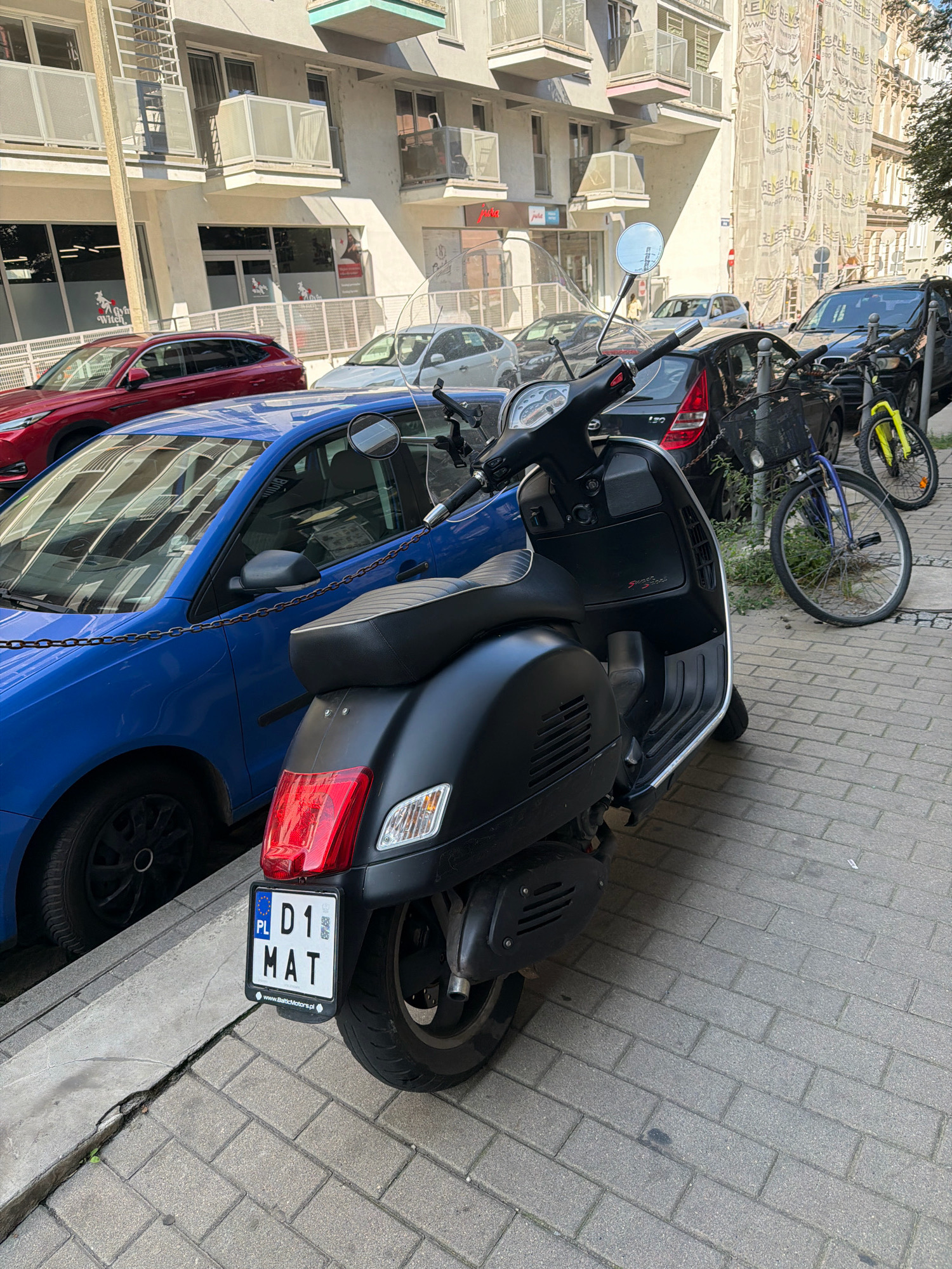 A black Vespa scooter is parked on a cobblestone sidewalk in front of a modern building in Wrocław, Poland. The scooter has a license plate that reads "PL D1 MAT". A blue car is parked next to the scooter, and other cars and bicycles are visible in the background. The building appears to be an apartment building with balconies.