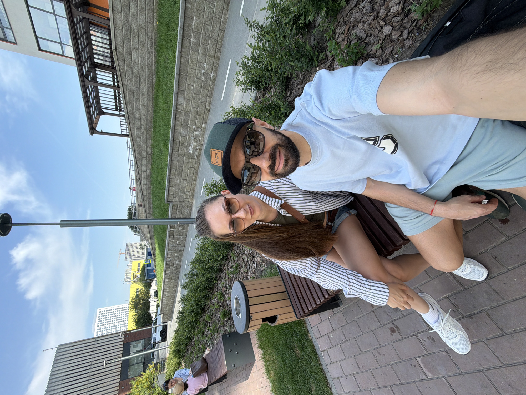The image depicts a man and a woman sitting closely on a wooden bench. The man has a beard, wears a hat and sunglasses, and a light blue T-shirt and shorts. The woman wears sunglasses, a striped shirt, and denim shorts. The background includes a brick walkway, greenery, buildings, and a blue sky with clouds. The image was taken outdoors in what appears to be a city park or public area in Tyumen, Russia.