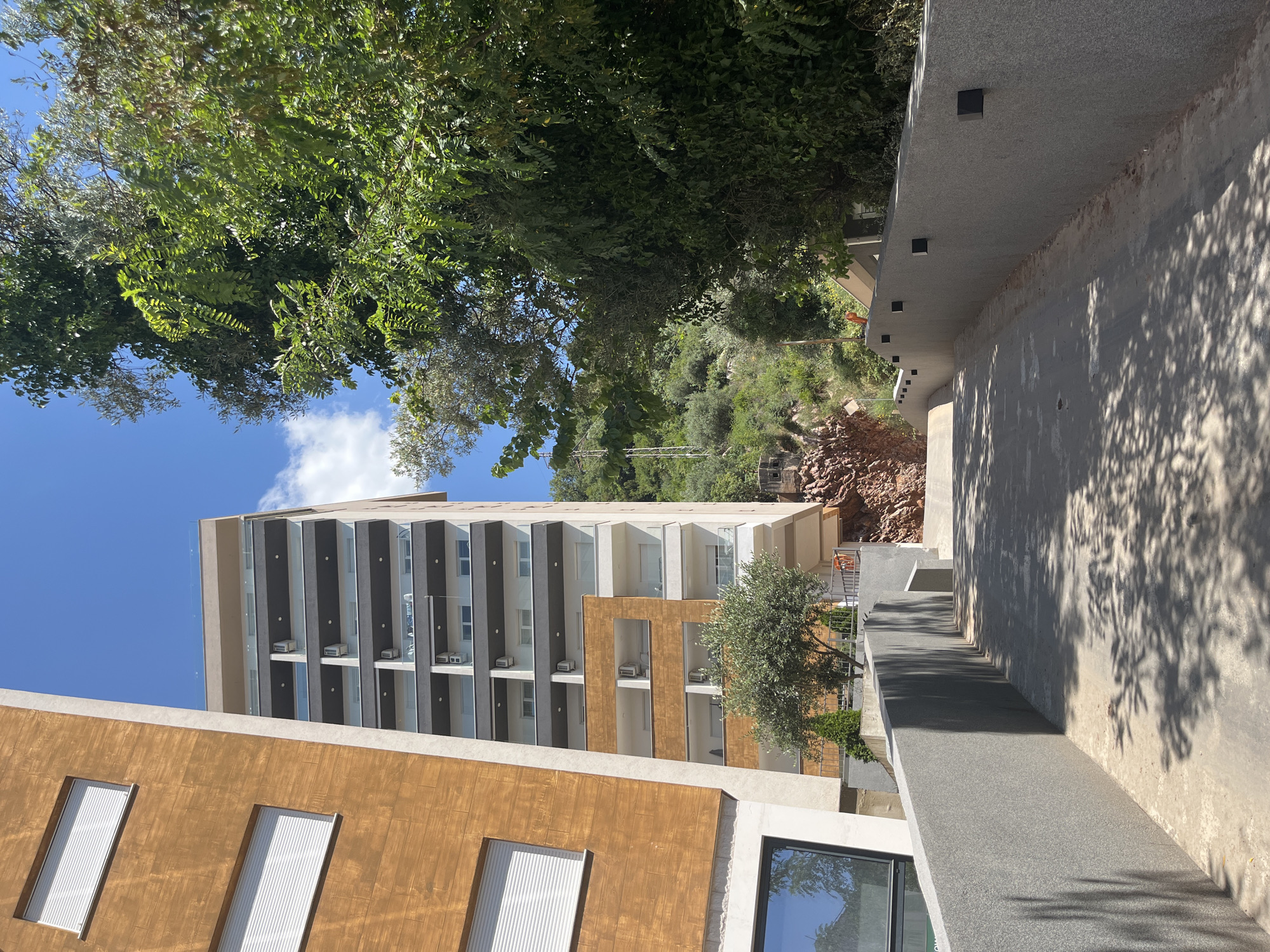 The image depicts a modern building complex under a clear blue sky with scattered clouds, located in Budva, Montenegro. The foreground features a paved pathway with grey concrete walls on either side, casting shadows from overhead foliage. The pathway slopes upwards, leading towards the buildings.
To the left, a building with a distinctive warm-toned, wood-like facade is visible, featuring horizontal elements and white-slatted windows. Adjacent to it, a taller, multi-story building presents a modern architectural style with dark grey and white accents, balconies with glass railings, and visible air conditioning units.
In the mid-ground, a section of the building complex reveals an exposed rock face and an olive tree. Beyond the buildings, a lush green hillside is covered in dense vegetation, with glimpses of what appear to be power lines and structures on the slope. The overall scene conveys a sense of contemporary construction integrated into a natural, hilly environment. No people or animals are visible in the image.