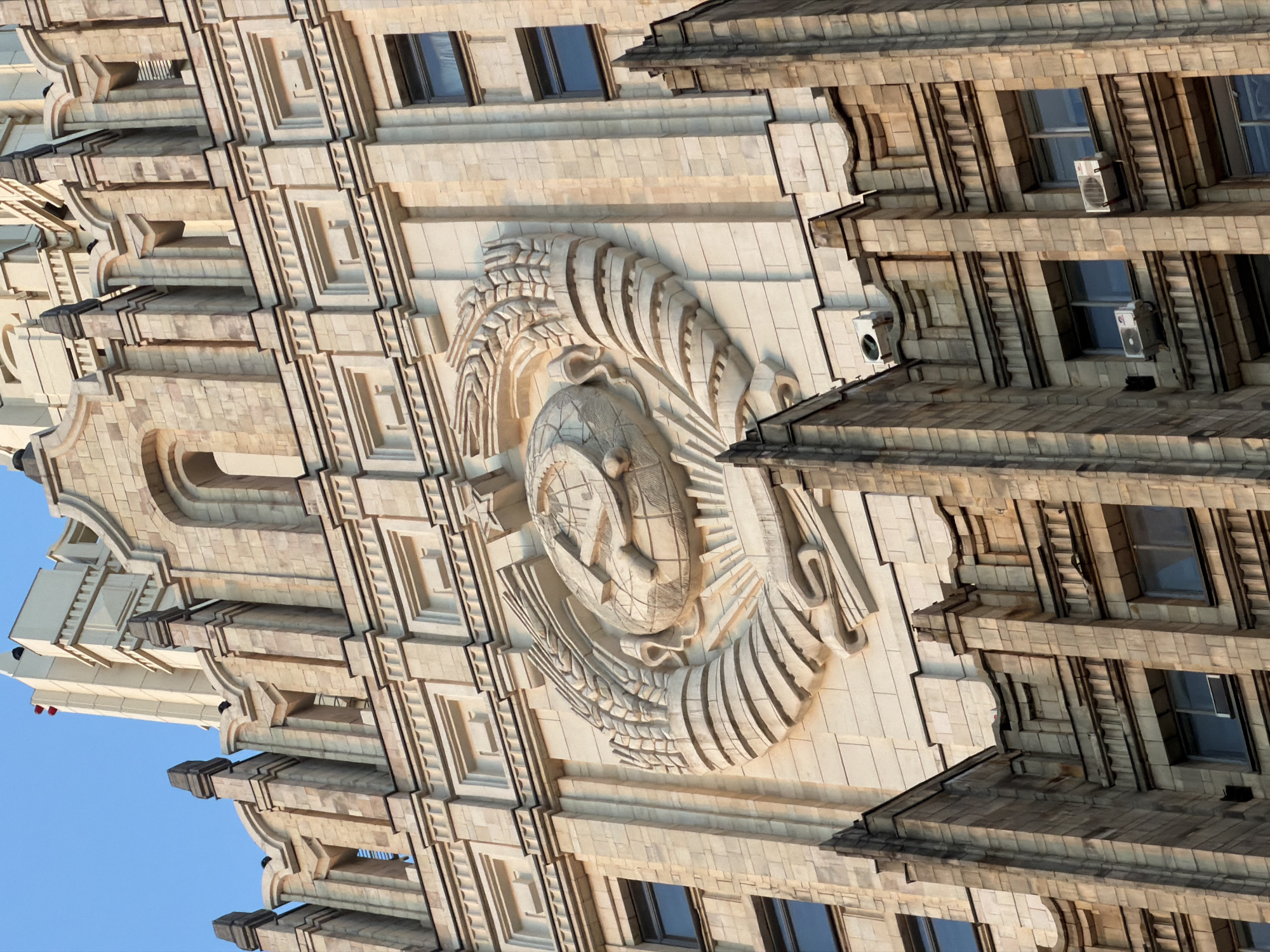 The image depicts a section of a building in Moscow, Russia, showcasing Soviet-era architecture. The building is light-colored, and features decorative elements. A prominent sculptural relief of the Soviet Union's coat of arms—a hammer and sickle overlaid on a globe and surrounded by wheat stalks—is centrally positioned on the facade. Below the Soviet coat of arms are the windows of the building. Air-conditioning units are affixed to some of the windows. The building's architectural style incorporates layered, stepped levels. The sky, a clear blue, is visible in the bottom left corner of the image.