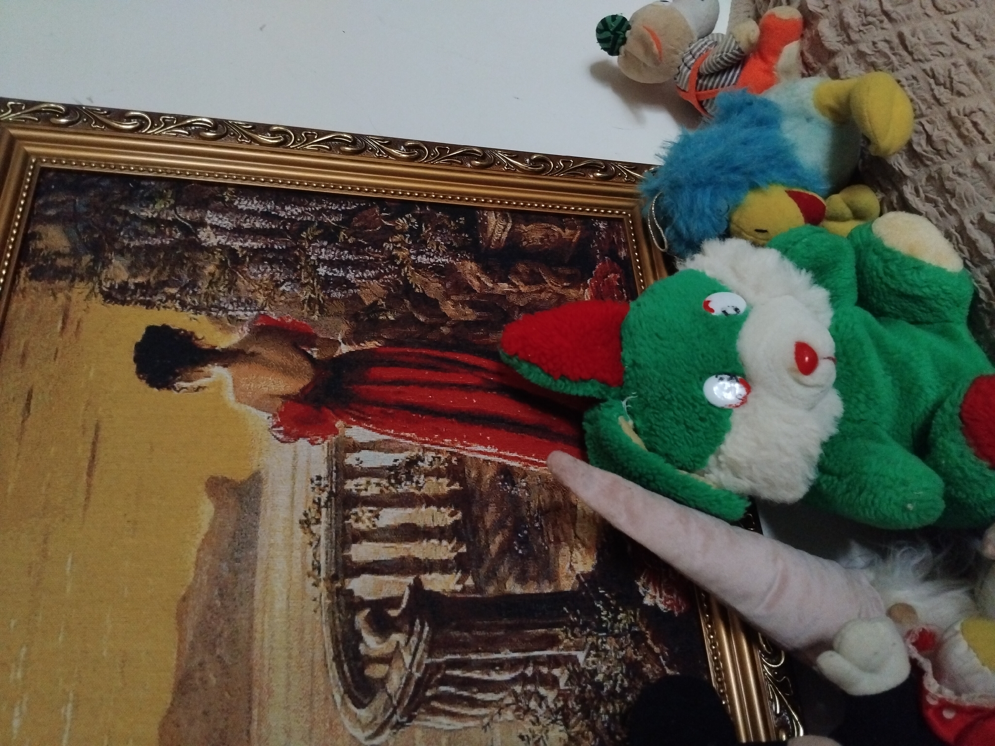 The image shows a framed painting of a woman in a red dress standing on a balcony overlooking a body of water and cliffs. In front of the painting are several plush toys, including a green bunny, a gnome, and a beige stuffed animal wearing an orange and gray striped shirt and orange pants. The plush toys are arranged on a textured surface that appears to be a blanket or bedspread. The painting is ornate with a gold-colored frame. The overall scene suggests an indoor setting, possibly a bedroom or living area. There is no discernible information in the image to indicate a specific location in the city of Akhaltsikhe, Georgia.