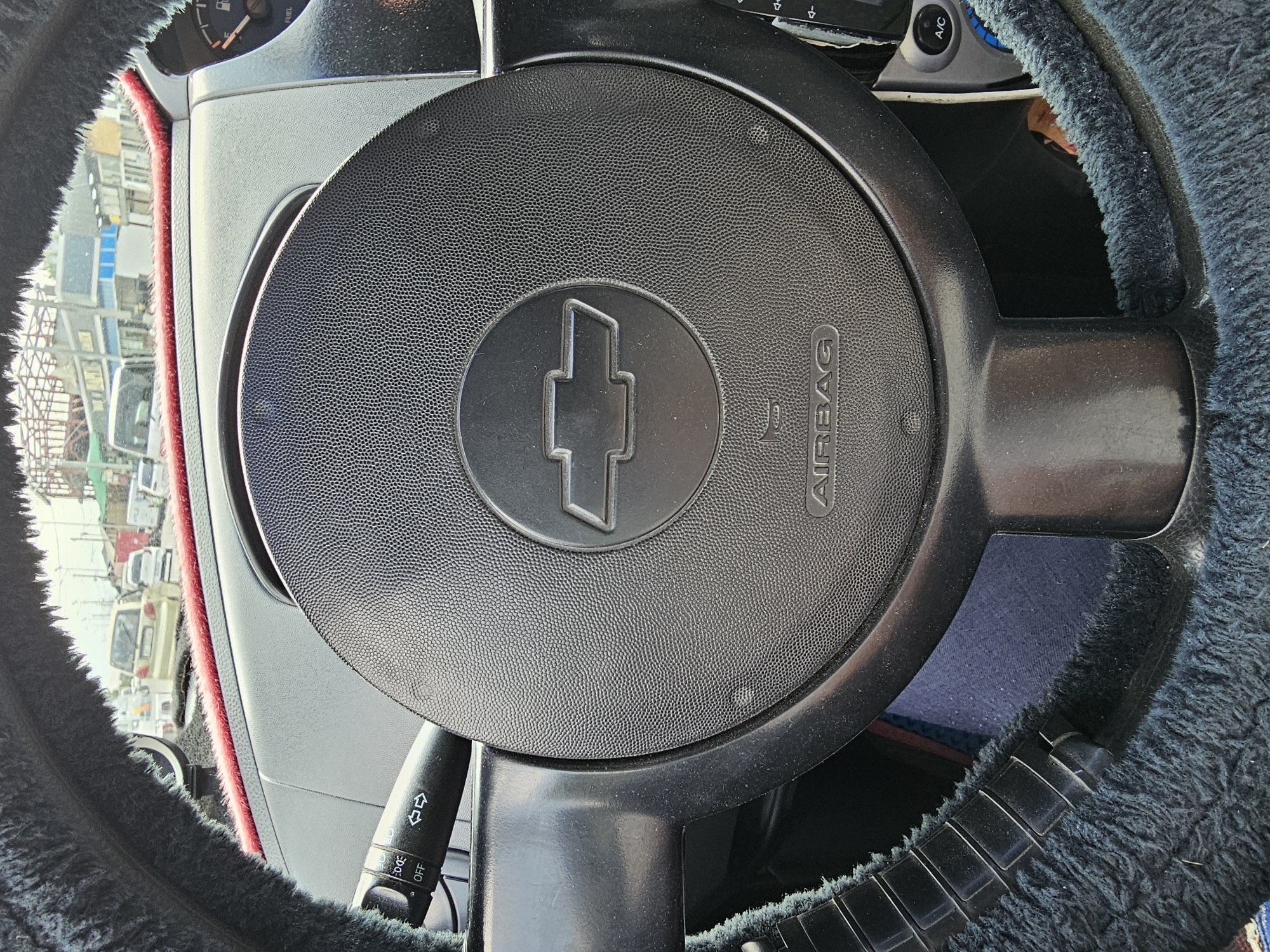 The image shows the interior of a car, likely parked or in slow-moving traffic. The focus is on the steering wheel, which has a black textured surface with a Chevrolet emblem in the center. The word "AIRBAG" is visible below the emblem. To the left of the steering wheel is a turn signal lever with markings for "OFF" and turn signals, as well as a light control with "AUTO" and "ON" settings. The steering wheel is covered in a black fluffy material with red trim. In the background, through the windshield, several vehicles are visible, suggesting an urban environment. Buildings are also present, indicating a city setting. The overall impression is that of a typical car interior in a traffic situation. There is no indication of the specific location of Özgön, Kyrgyzstan.