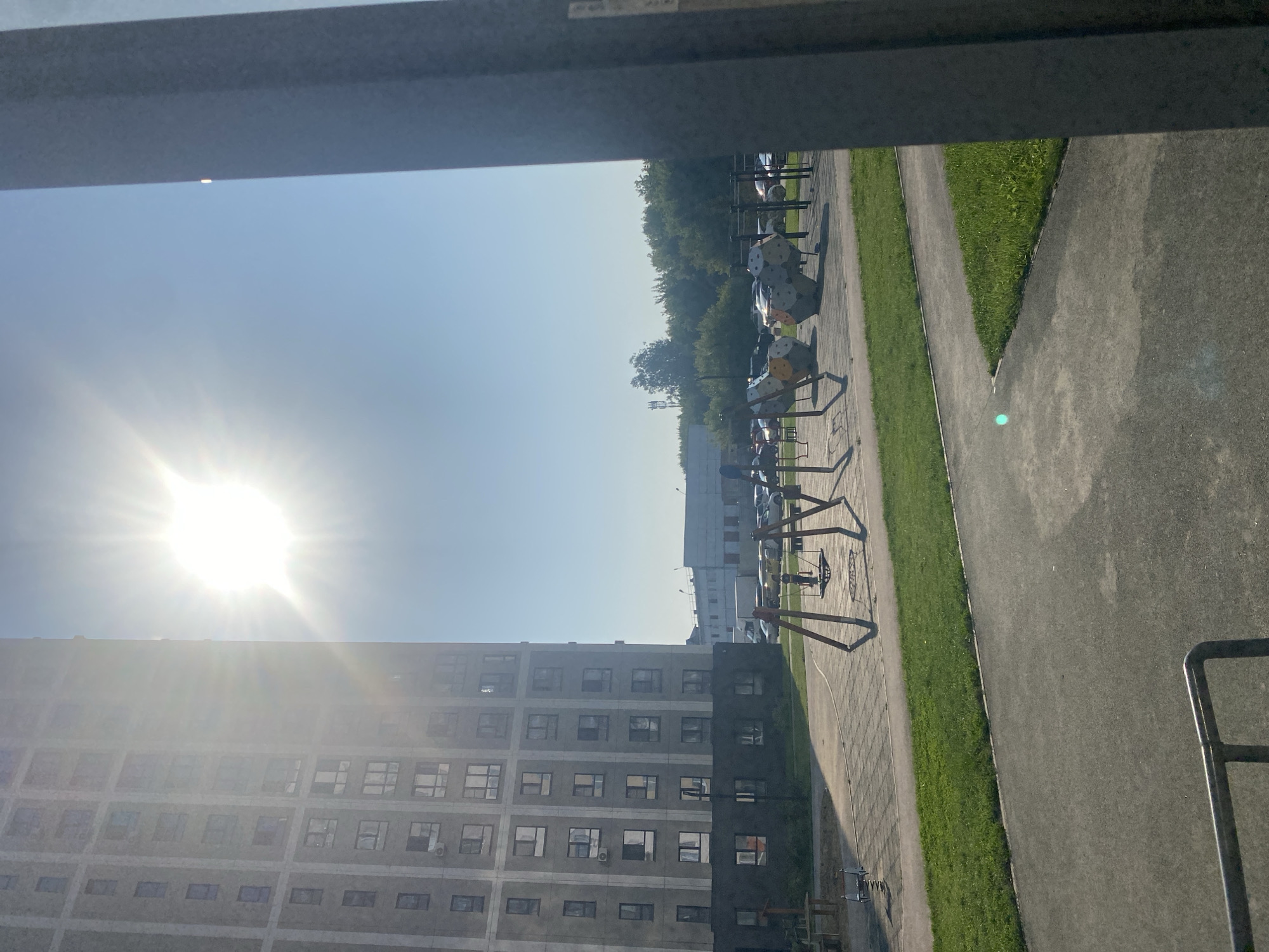 A bright sun with visible rays shines in a clear blue sky, partially obscured by a tall, modern apartment building to the left. The building's facade is light grey with numerous windows. In the foreground, a playground area is visible, featuring swings and abstract climbing structures. Beyond the playground, a parking lot is filled with cars, and further in the background, trees and other buildings are present. The scene appears to be a residential area in Novosibirsk, Russia, likely during daytime.