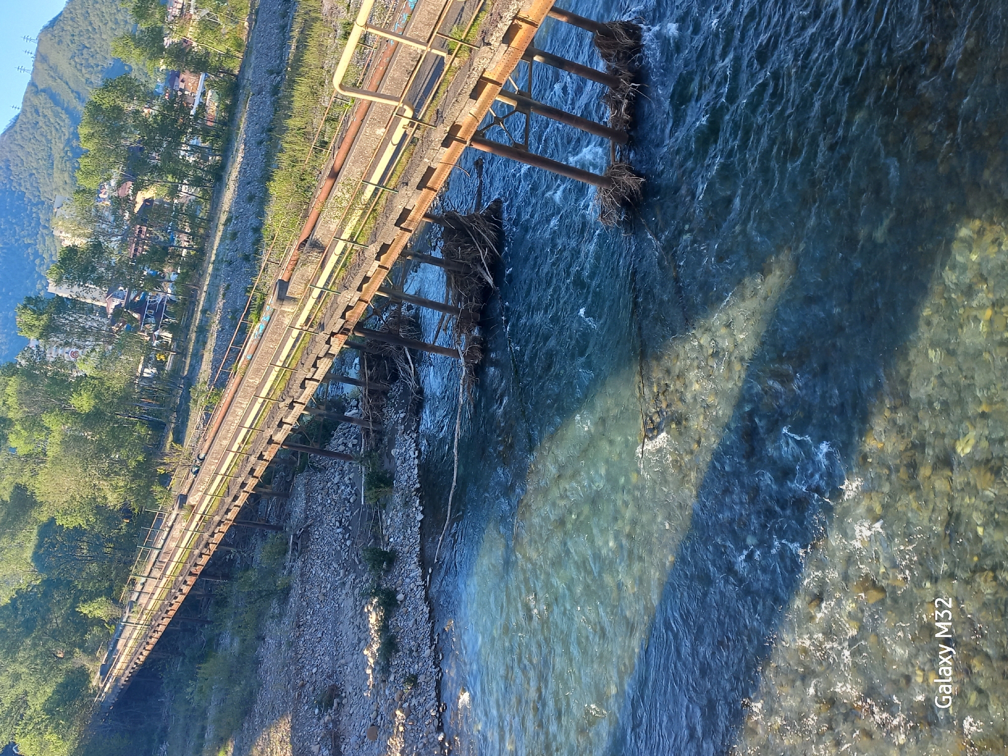 An elevated railway bridge spans a clear river in Sochi, Russia. The bridge is rusty, supported by metal columns in the water, some with accumulated debris. The riverbed is visible through the water. One bank is rocky and partially vegetated, while the opposite bank features a line of trees and buildings. Mountains are visible in the background. The photo appears to be taken from a high angle.