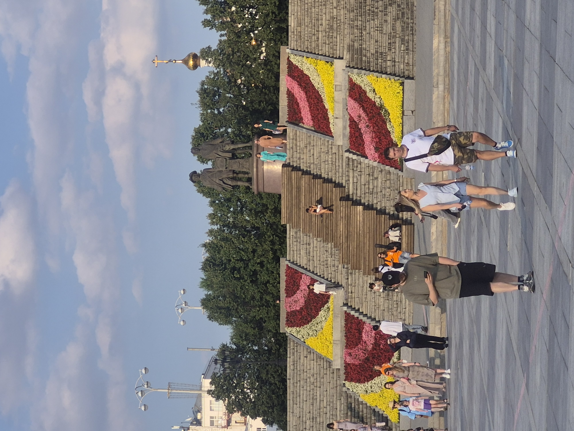 The image depicts a sunny day in Yekaterinburg, Russia, likely in an open public space with a large, tiered set of stone stairs. Two prominent statues of men stand at the top of the stairs, and a golden-domed orthodox church with a cross is visible in the background. The stairs themselves are decorated with large, colorful flower arrangements in red, pink, and yellow, forming diagonal stripes. Several people are walking on the plaza in the foreground and ascending the stairs. A man in a green t-shirt and black shorts is looking at his phone in the foreground. To his right, a couple is walking, with the man wearing a white shirt with a logo and camouflage shorts, and the woman in a light blue top and denim shorts. Several other individuals and groups are scattered throughout the scene, engaged in walking or sitting. The environment appears to be a well-maintained urban area, possibly a park or central square, with clear blue skies and scattered clouds.