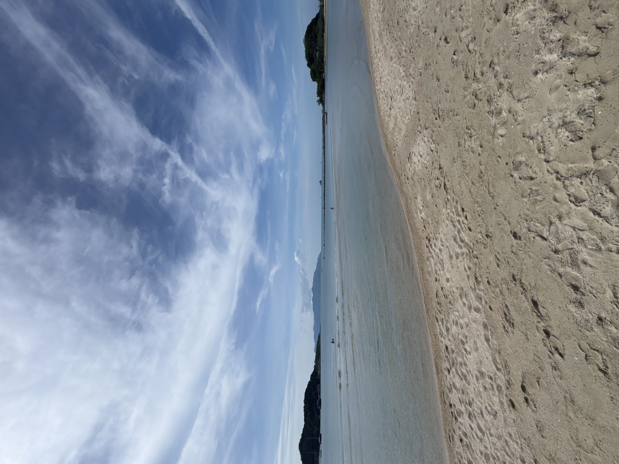 A beach scene on a clear day, with a sandy foreground and a calm body of water leading to distant islands under a sky with scattered clouds. In the foreground, footprints are visible in the sand. The water is shallow and transparent, with faint ripples. A few small boats and people are present in the water and along the distant shoreline. The overall impression is one of a serene coastal landscape.