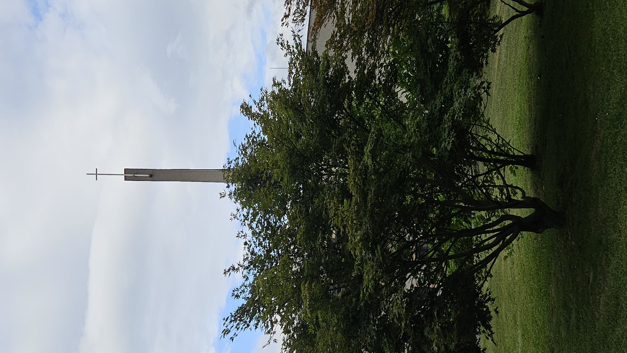 The image captures a view of a cross atop a tall, slender concrete structure, possibly a church steeple or bell tower, against a backdrop of a cloudy sky. In the foreground, lush green grass carpets the ground, with several leafy trees providing a natural frame. To the right, a portion of a building with a light-colored facade is visible behind the trees. The overall scene suggests an outdoor setting, likely in Prešov, Slovakia, given the context. The time of day appears to be daytime, with diffused lighting due to the cloudy conditions. No people or specific actions are depicted in the image.