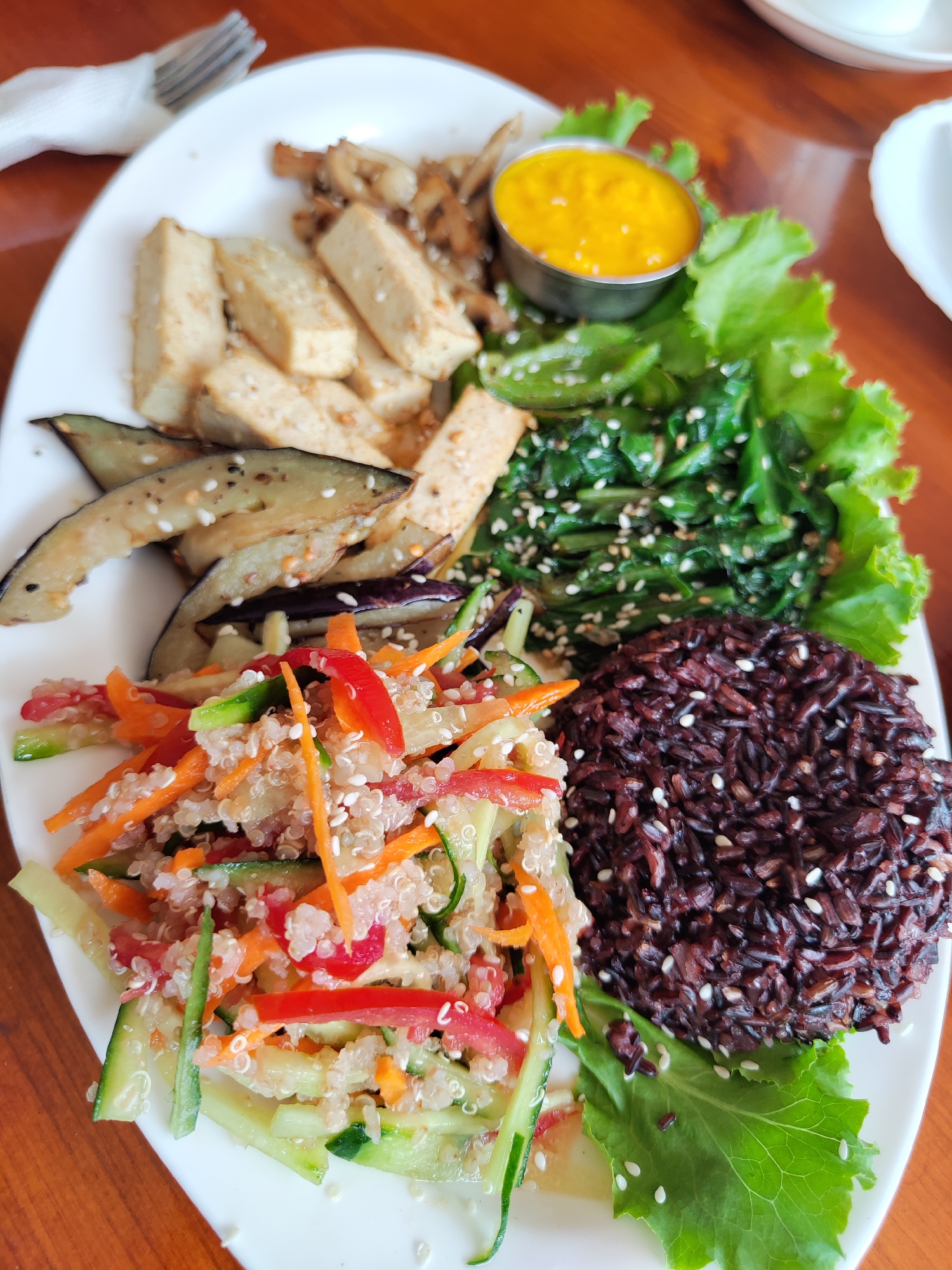 A high-angle, close-up shot presents a white oval plate filled with an assortment of foods, likely in a restaurant located in Bhagsu, India. The plate is arranged with sections of fried tofu, sautéed mushrooms, fried eggplant, a quinoa salad with carrot, cucumber, and bell pepper, dark rice, and a leafy green vegetable. There is a small metal ramekin filled with a yellow sauce positioned near the top right of the plate. The entire dish is sprinkled with sesame seeds. In the upper left corner of the frame is a fork resting on a napkin. A wooden table provides the background. A small portion of a white dish is visible in the upper right corner.