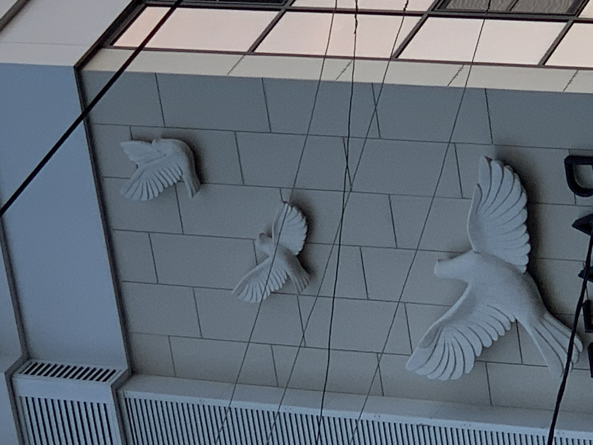 A facade of a building features three white dove sculptures, each with outstretched wings. The doves are positioned at different heights and angles. The wall behind them is covered in light gray square tiles. Thin black wires crisscross the image in front of the wall and sculptures. The top left of the image shows a section of the building with horizontal white louvers, likely part of an air vent. To the right, a portion of a modern building with reflective windows is visible. The lighting suggests it is either early morning or late afternoon. The scene is devoid of any people or animals.