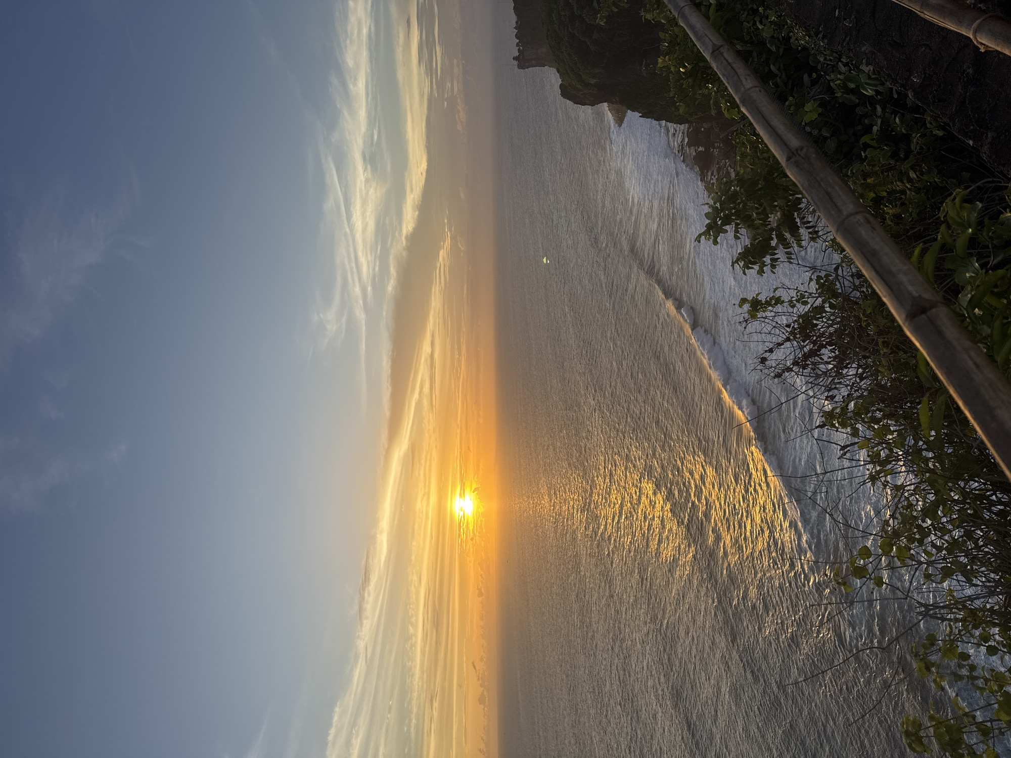 The image shows a sunset over the ocean. The sun is setting on the horizon, casting a golden glow on the water. The sky is blue with wisps of clouds. The coastline has cliffs and vegetation. A portion of a bamboo structure is visible in the foreground. The location appears to be a high vantage point overlooking the ocean.