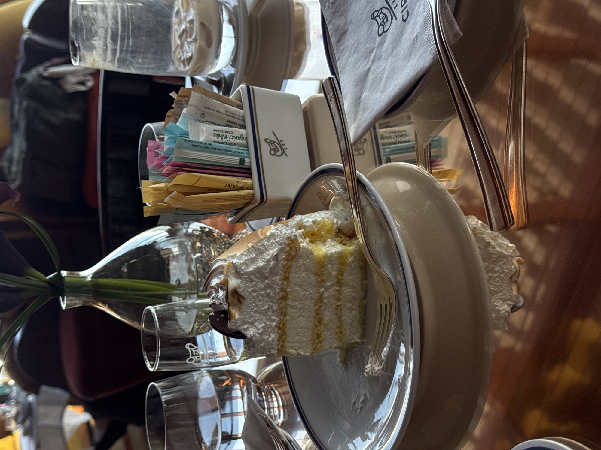 The image captures a close-up view of a dining table inside a Cipriani restaurant or cafe in New York City, United States of America. No individuals are present.
Dominating the foreground is a partially consumed slice of layered white cake, featuring white cream or frosting between yellow cake layers, topped with a dark, perhaps chocolate, embellishment and a hint of torched meringue. It rests on a white plate with a blue rim. A gold-colored fork, bearing cake residue, lies on the plate.
To the left of the cake, two clear glass tumblers are visible; one contains water, the other appears empty. To the right, a clear glass holds remnants of an iced beverage, with melted ice cubes and a small amount of milky liquid. A small, white rectangular ceramic caddy, detailed with blue stripes and a stylized bear logo, sits nearby, filled with various single-serve sweetener packets, including "Organic White Cane Sugar." Below this, a folded white napkin, displaying the "CIPRIANI" logo and the same stylized bear emblem, is visible.
In the mid-ground, a clear glass vase containing dark purple, slender-petaled flowers with green stems serves as a centerpiece. The table surface is polished and highly reflective, mirroring the objects and ambient light. The background is softly blurred, showing glimpses of dark upholstered furniture and a warm, interior lighting scheme, consistent with an upscale dining establishment. The visible Cipriani branding on the napkin and caddy confirms the location.