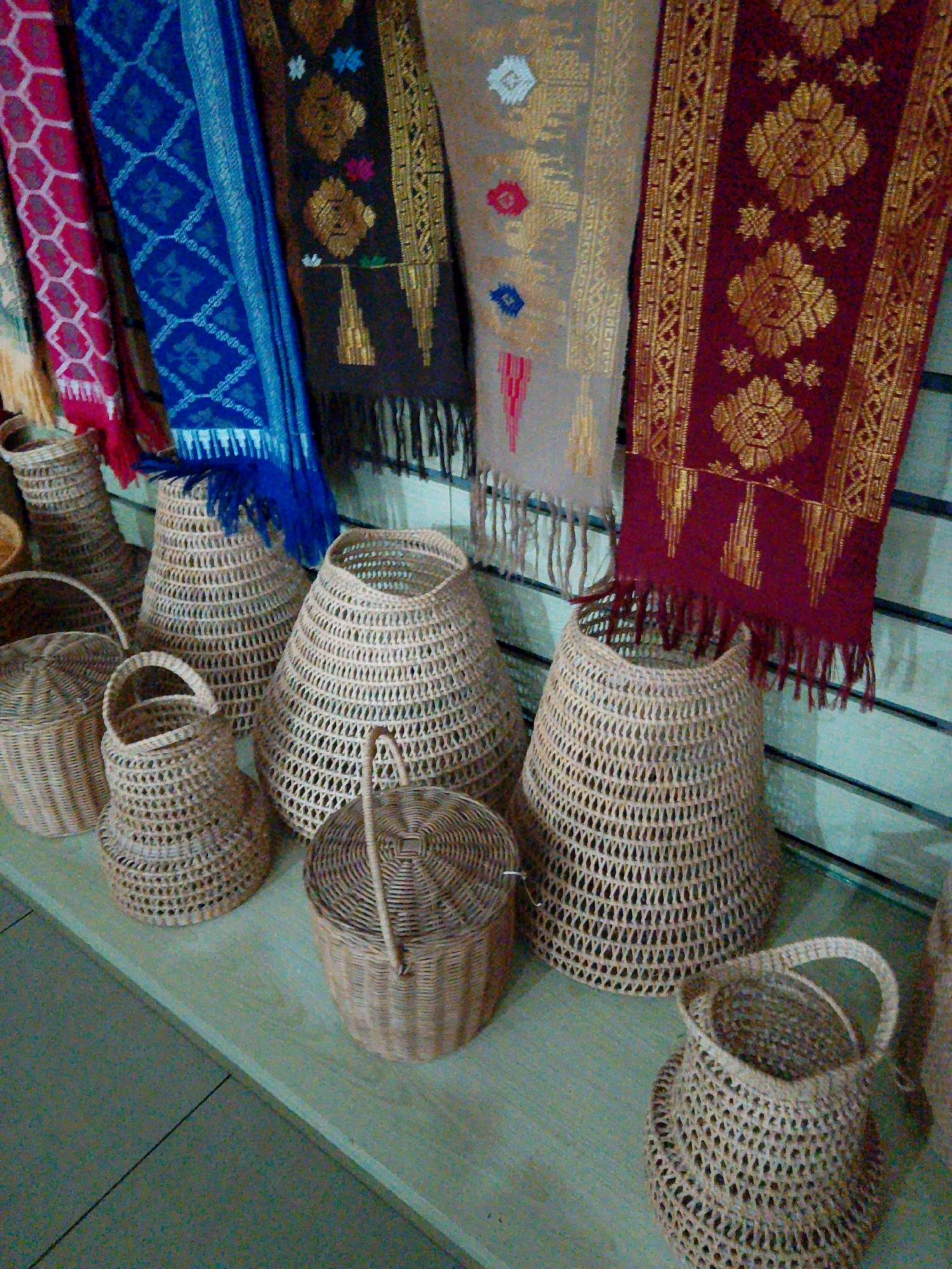 The image displays a collection of woven baskets and intricately patterned textiles, likely in a market or artisan shop in Bunkate, Indonesia. The woven baskets are of various sizes and shapes, with open, lattice-like designs, suggesting they are made from natural materials like rattan or bamboo. Some baskets have handles, while others are cylindrical or conical. The textiles, hanging vertically behind the baskets, feature rich colors and detailed patterns, possibly representing traditional Indonesian designs. They appear to be scarves or sarongs, with fringed edges. The overall scene suggests a vibrant display of local crafts.