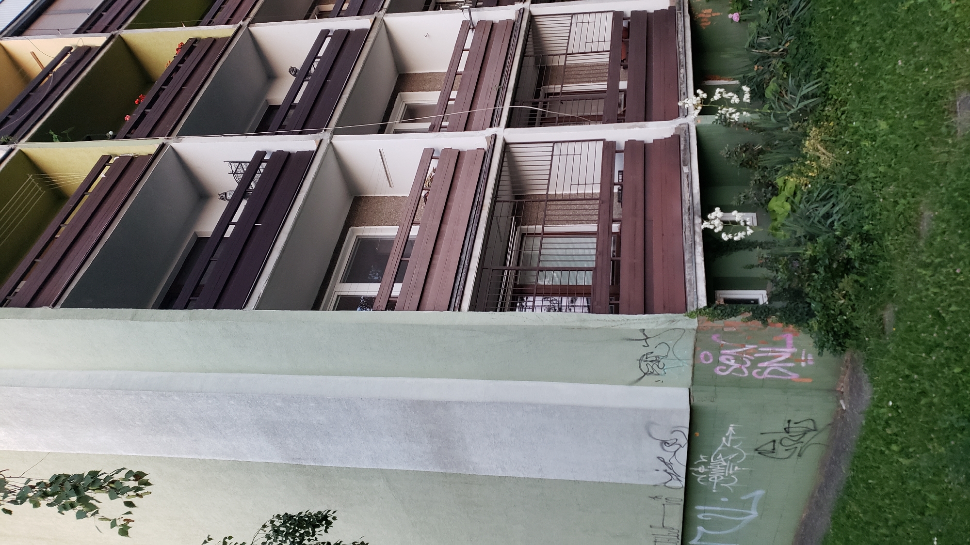 The image shows a close-up, low-angle view of a multi-story apartment building with balconies in Prešov, Slovakia. The building has a pale green exterior wall on the left, which is covered in graffiti. The right side of the building features multiple levels of balconies with dark brown railings and supports. Some balconies have windows and appear to have potted plants. The lower level of the building has a garden area with various plants and flowers, including white blossoms. The scene is observed during daylight, with a grassy area in the foreground.