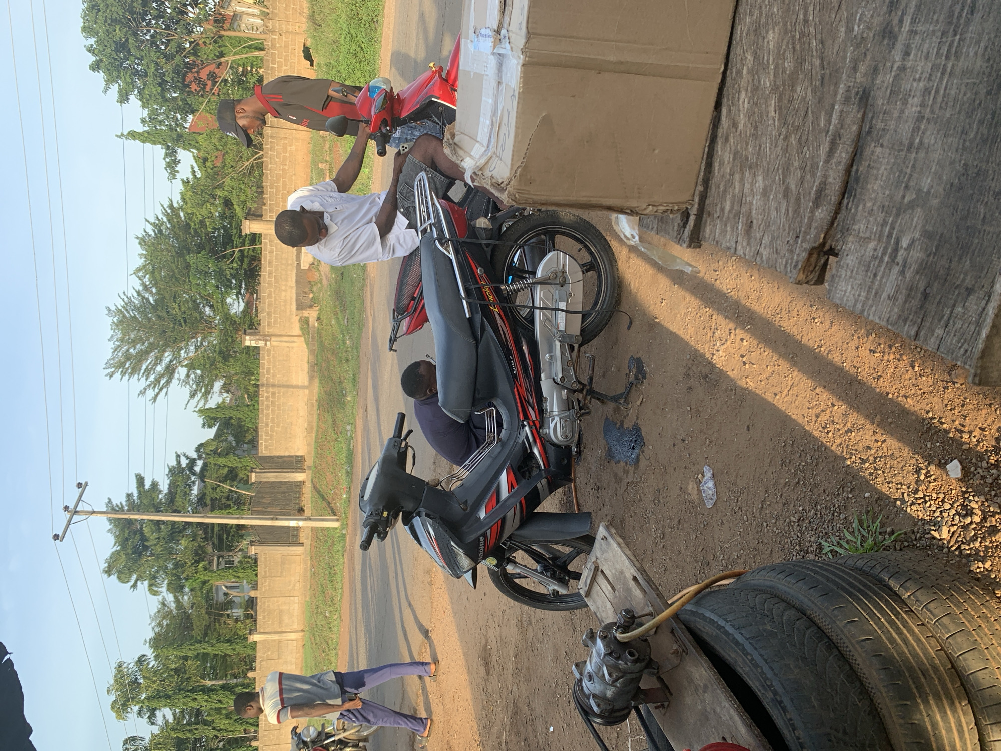 Two men are working on a red and black Haojue motorcycle in an outdoor setting. One man is seated on a red motorcycle while the other, wearing a white shirt, is attending to the Haojue motorcycle. An older man is seated behind him on the Haojue motorcycle, seemingly assisting. The motorcycle appears to be undergoing some maintenance or repair, as indicated by the dark liquid on the ground beneath it. A stack of tires and an air compressor are present near the motorcycle, suggesting a repair shop or roadside service area. The environment includes a dirt road or shoulder, a wooden plank serving as a makeshift work surface, and a large cardboard box. In the background, there is a paved road, a partial view of a residential area, trees, and an electrical pole, which helps establish a location in Kabba, Nigeria.