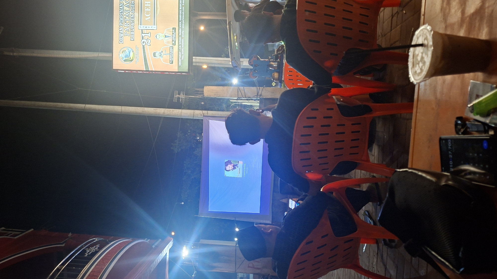 In Keudeteupinpunti, Indonesia, at night, an outdoor screening is taking place. The focus is on a large projected image displaying a profile picture and text. Three individuals are seated in orange plastic chairs, facing the screen. The person in the leftmost chair wears a black cap and is looking at a smartphone. The other two individuals are also facing the screen, with one appearing to be engaged with a smartphone. In the foreground, a table holds a black bag, a calculator, a lighter, a pack of cigarettes, and a cup of iced coffee with a straw. Above and to the right, a billboard for "DEWAN PIMPINAN PARTAI NASIONAL ACEH KABUPATEN ACEH" is visible, featuring portraits of two men and text in Indonesian. Power lines and streetlights are also present in the environment. The scene suggests a community gathering or event in a public space.