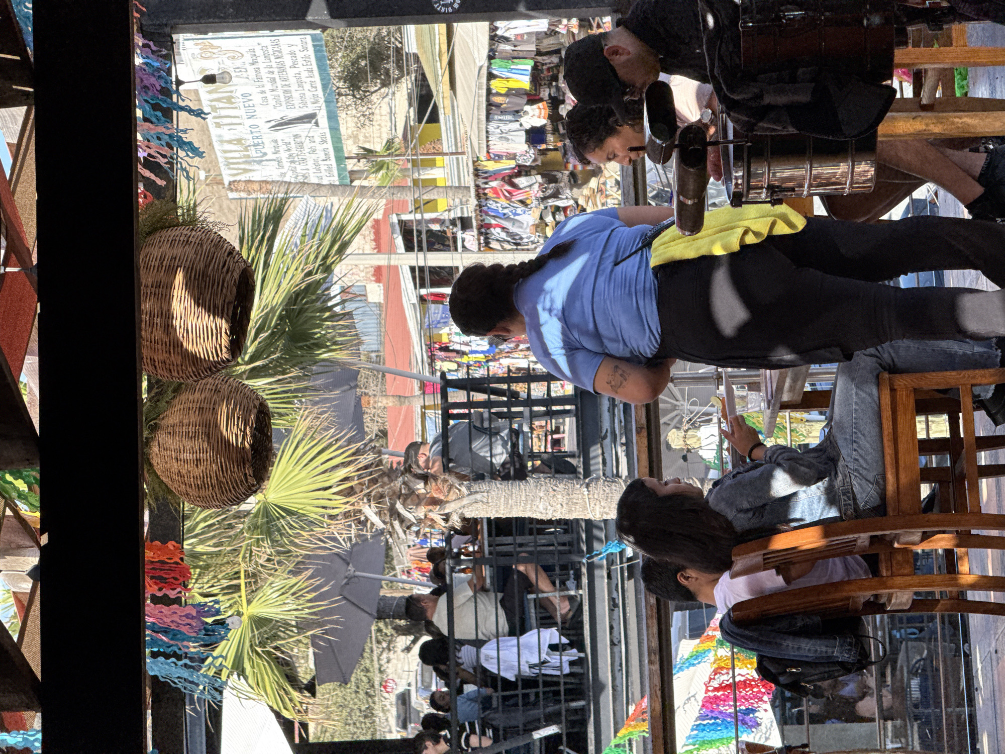 This image depicts a scene at Villa Titan, Puerto Nuevo, Mexico. A group of people are seated at tables under an outdoor canopy, with palm trees and a marketplace visible in the background. In the foreground, a woman in a blue shirt and black pants stands, wearing a yellow towel over her shoulder. She is facing away from the camera, towards a table where two people are seated. To the right, another woman in a denim jacket is seated at a table, holding a plate. Men are also visible at tables, some wearing hats and sunglasses. Colorful banners and woven baskets hang from the canopy. The marketplace in the background features racks of clothing and other merchandise.