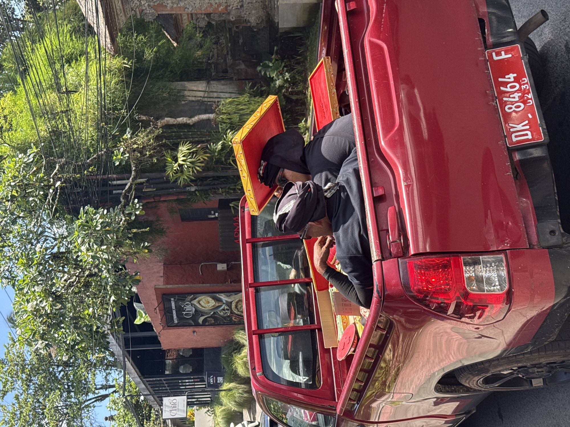 A red pickup truck is parked on a sunlit street, facing away from the viewer, with its tailgate down. The image is oriented vertically (rotated 90 degrees clockwise). In the open bed of the truck, two individuals, clad in dark clothing and head coverings, are seated as passengers. They are positioned amidst several elaborately decorated items, which appear to be gold-trimmed wooden trays with red bases, possibly ceremonial offerings. One such tray is prominent near the person closer to the camera.
The truck's red license plate, "DK 8464 F," is clearly visible on the tailgate, with "02 30" inscribed below, likely indicating an expiration date of February 2030. The "DK" prefix confirms the vehicle's registration in Bali, Indonesia, consistent with the Ubud District context. The truck also features red taillights and a spare tire mounted beneath the rear bumper.
The background is rich with lush, green tropical vegetation, including dense foliage and trees. Numerous black electrical or communication wires are strung overhead, crisscrossing the scene. Buildings with distinctive red-painted walls and dark windows line the street. One building prominently displays multiple signs for a business named "Chiiô," with additional text on one sign reading "GASTRONOMIC JOURNEY" and another small sign indicating "OPEN."
The bright, clear daylight suggests it is late morning or afternoon. The overall scene depicts a moment of daily life or transport in a tropical setting, possibly involving cultural items given the nature of the decorative trays and the Indonesian location. No specific emotions are discernible from the individuals in the truck.