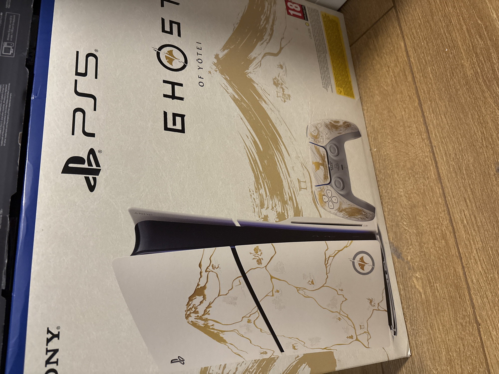 This image displays the packaging for a PlayStation 5 (PS5) console, featuring a custom "Ghost of Yotei" design. The box is predominantly white with gold and black graphics and text, positioned on a light brown wooden floor.
The main subjects depicted on the box are a PS5 console and a DualSense controller, both showcasing a unique white and gold aesthetic. The console, shown horizontally, features an intricate gold map-like pattern with mountain silhouettes across its white side plates. The DualSense controller, positioned above the console image, also has a matching white and gold custom design.
Visible text on the packaging includes the "PS5" logo, the "GHOST" title with a circular emblem (resembling a ginkgo leaf) within the "O," and "OF YÖTEI" printed below it. The "SONY" logo is partially visible at the bottom left. An age rating of "18" within a red circle is present on the top right. Various unreadable small print and legal disclaimers are also visible on the box.
No people are present, and no specific activity is taking place; it's a static shot of a product box. The setting is indoors, with the wooden floor suggesting a domestic environment.