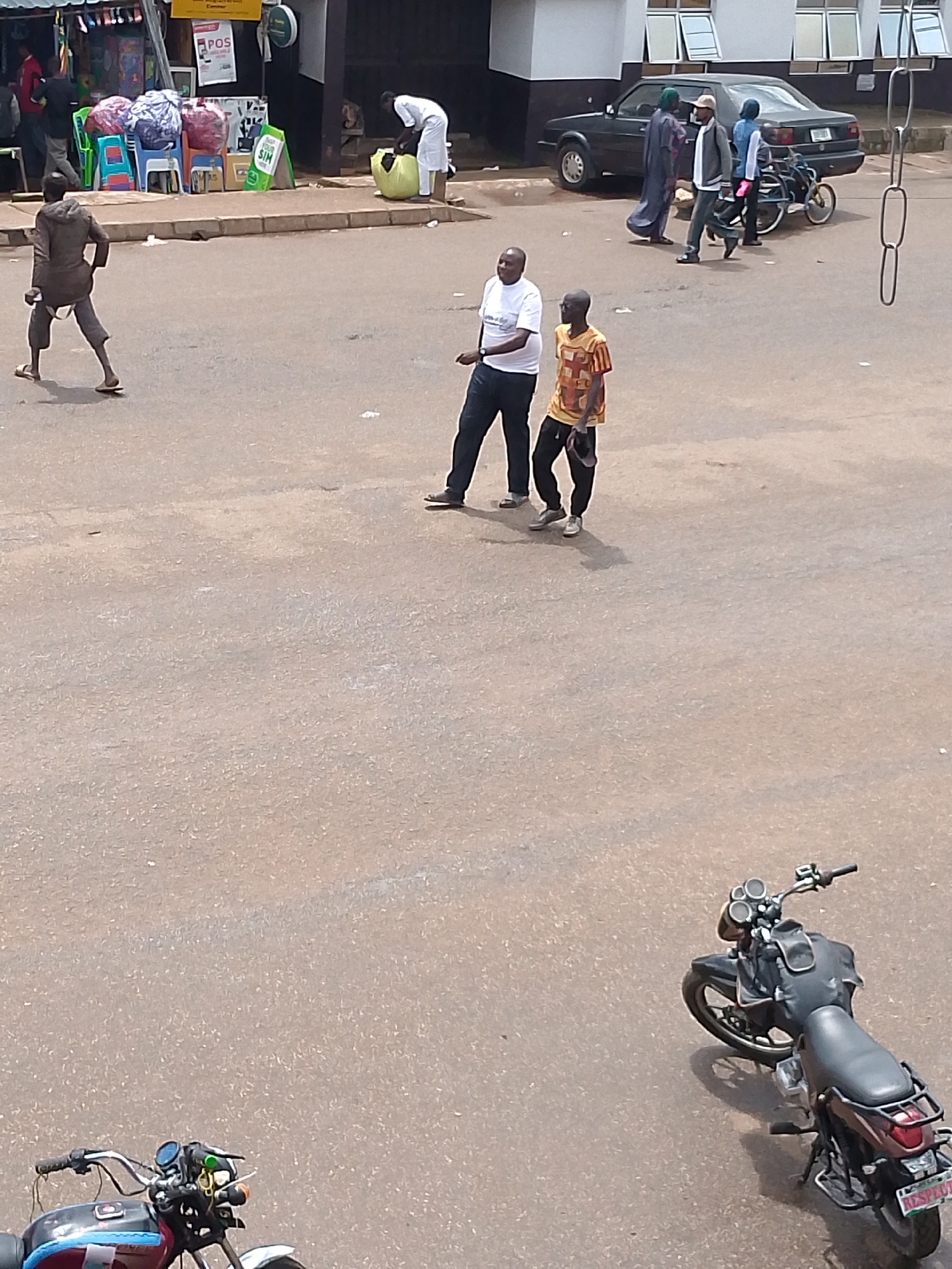 In Bukuru, Nigeria, two men are engaged in a playful interaction in the street. One man, wearing a white t-shirt and dark pants, stands with his arms extended, while the other, in a yellow and red patterned shirt and dark pants, appears to be mimicking his posture. Nearby, a third man in a grey tracksuit walks away from the viewer. In the background, a dark sedan is parked, and several people are seen near a market stall with colorful bags and chairs. A motorcycle is parked in the foreground to the right, with its license plate visible. The scene takes place on a paved street, likely a commercial area, with buildings and a market visible in the background.