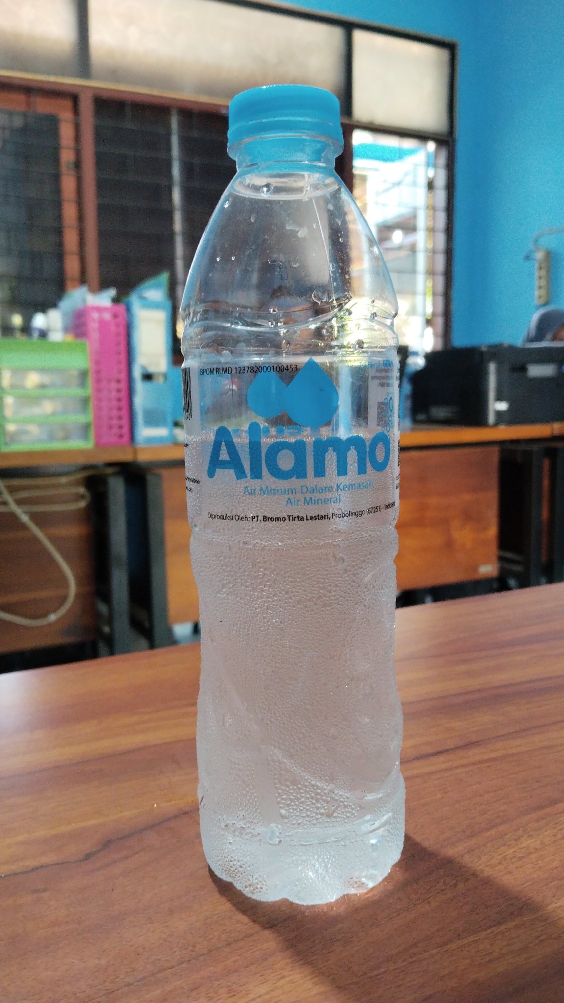 A close-up, eye-level, medium shot captures a condensation-covered plastic bottle of "Alamo" brand mineral water. The bottle stands upright on a wooden table, its surface slick with droplets of water. The bottle has a blue cap and a label that features two blue water drop graphics above the brand name. The text on the label indicates the water is "Air Minum Dalam Kemasan Air Mineral," and it is produced by "PT. Bromo Tirta Lestari, Probolinggo (67251) - Indonesia." The background is blurred but suggests an indoor setting with shelves containing items, and office furniture. The overall lighting is bright, highlighting the condensation on the bottle. The image does not provide any visual cues that would allow for specific localization to Buduan, Indonesia, or any other specific city.