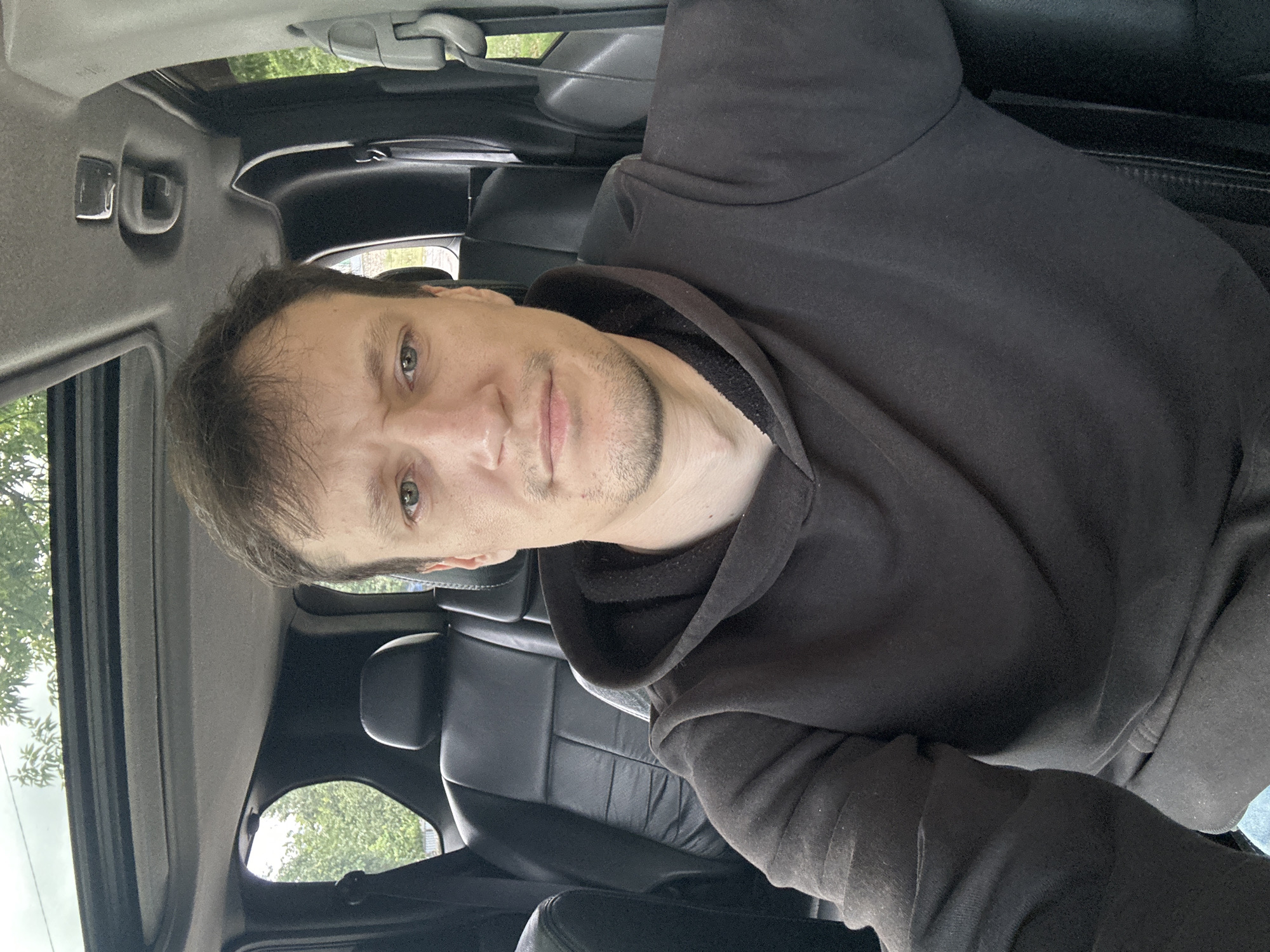 A man is seated in the driver's seat of a car, facing the camera. He is wearing a dark hooded sweatshirt. The interior of the car is visible, including black leather seats and a sunroof. Outside the car, trees and foliage can be seen. The setting appears to be a daytime outdoor environment, possibly in or near the city of Makiv, Ukraine, given the context provided.