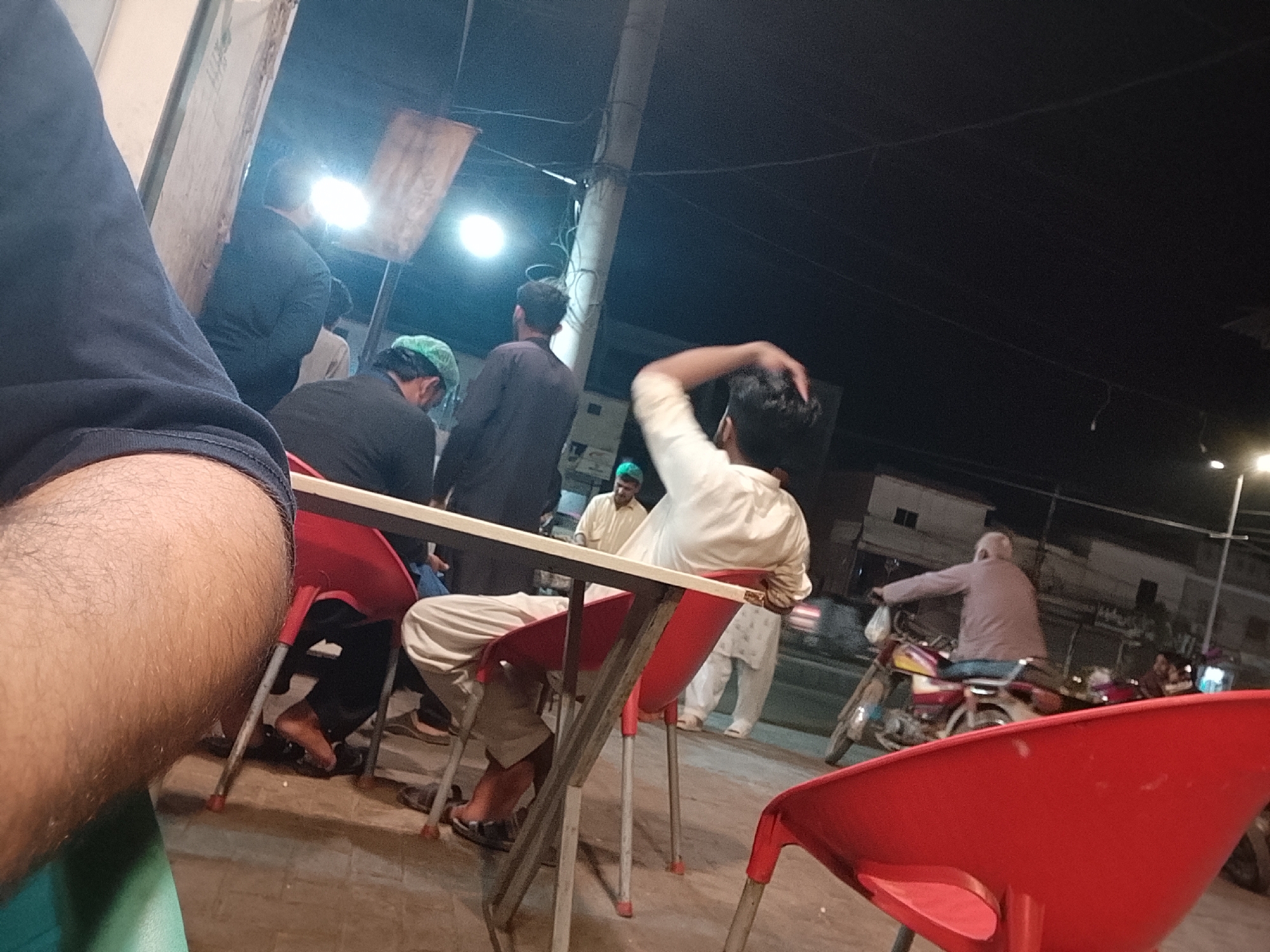 The image depicts an outdoor scene at night, likely a street-side eatery. Several men are present, some standing and others seated at tables with red plastic chairs. One man is seen rubbing the back of his head. Another man is riding a motorcycle past the group. Overhead, there is lighting illuminating the area. The location is believed to be in Multan, Pakistan, based on contextual information. The setting suggests a casual social gathering or a stop at a local food establishment. The viewpoint is that of a person seated at a table, with a portion of the person's leg and clothing visible in the foreground.