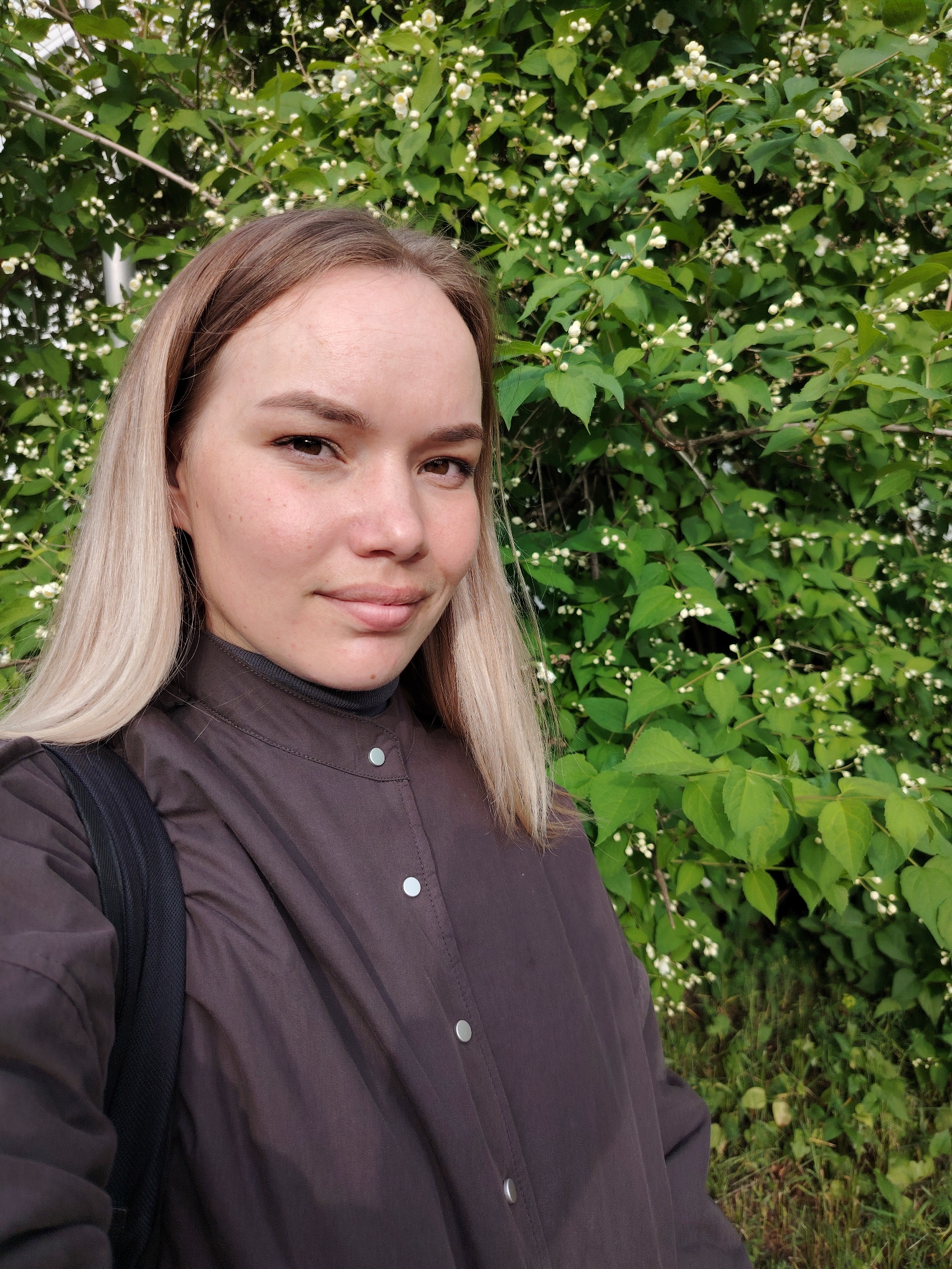 A woman with fair skin, brown eyes, and long, straight blonde hair with brown roots poses for a selfie. She has a neutral expression. She wears a brown, collared button-down jacket over a darker turtleneck sweater. A black backpack strap is visible over her left shoulder.
The background is a dense bush with green leaves and small white flowers. The photo was taken outdoors. The location is Chaikovsky, Russia.