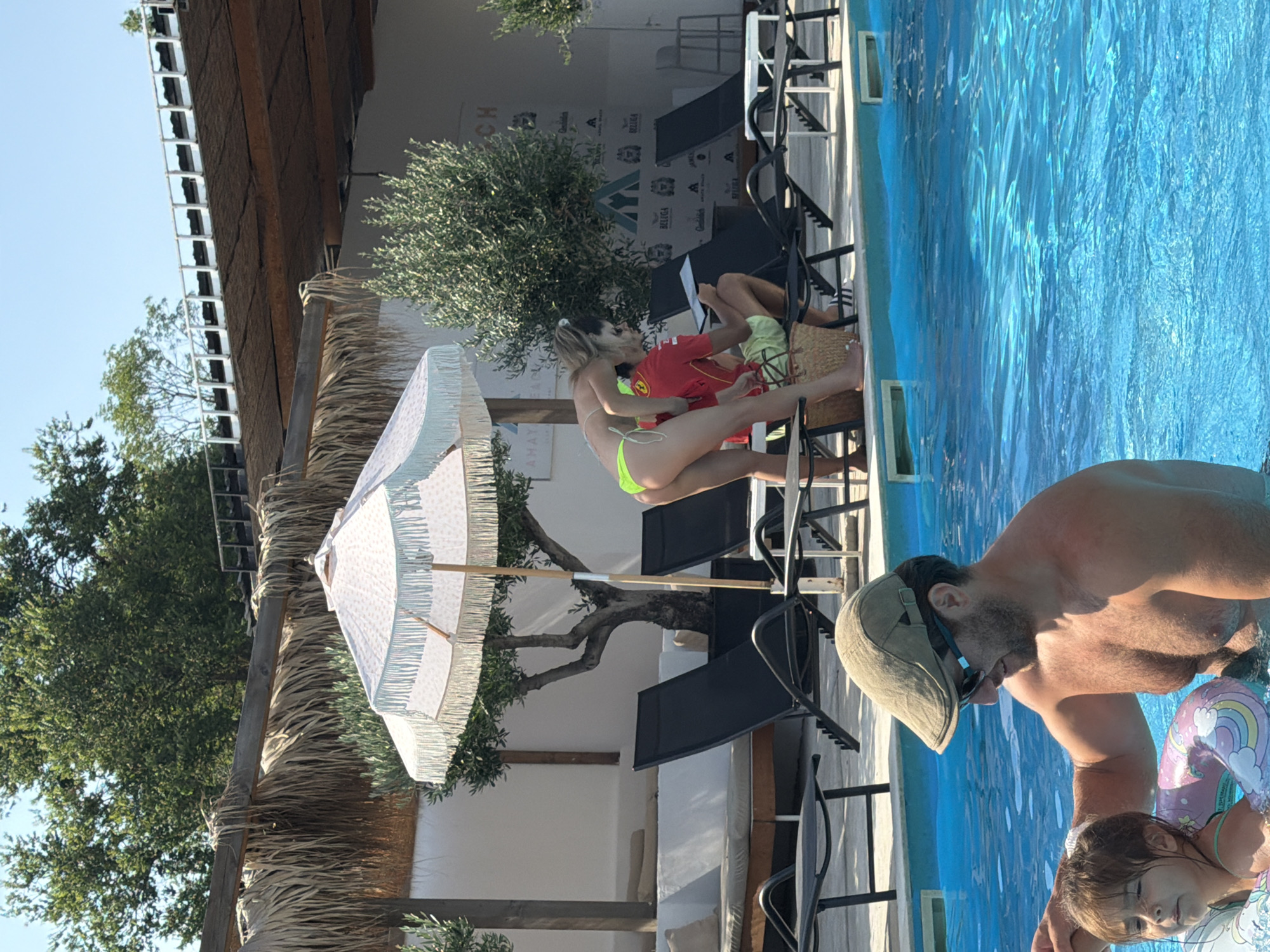 In a poolside setting in Burgas, Bulgaria, a man wearing a tan cap and sunglasses is in the bright blue water, looking down towards a young child. The child, wearing a colorful float with a rainbow design, is also in the water. In the background, a woman in a neon green bikini is sitting on a chair with a man wearing a red shirt, who is holding a piece of paper. A white umbrella with a fringed edge stands nearby, casting a shadow. The scene appears to be during daylight, with a clear sky. The overall atmosphere seems relaxed and summery. Visible text includes "BEACH" on a sign in the background.