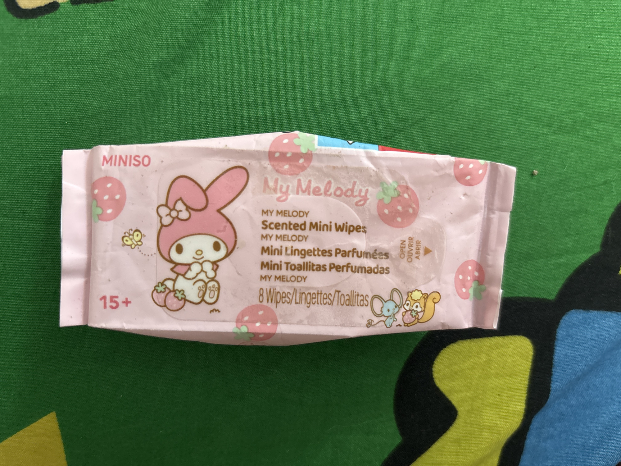 A pack of "My Melody Scented Mini Wipes" is centrally positioned on a green and blue patterned surface. The wipe pack features the My Melody character and strawberry illustrations, with text in English, French, and Spanish. The upper left corner has "MINISO" written on it, and the bottom left corner has the label "15+". The back of the package contains a red arrow symbol indicating the way to open the wipes, with the words, "OPEN, OUVRIR, ABRIR" written next to the symbol. The image suggests a domestic or retail environment, but there is no geographical context specifying its location as North Jakarta, Indonesia.