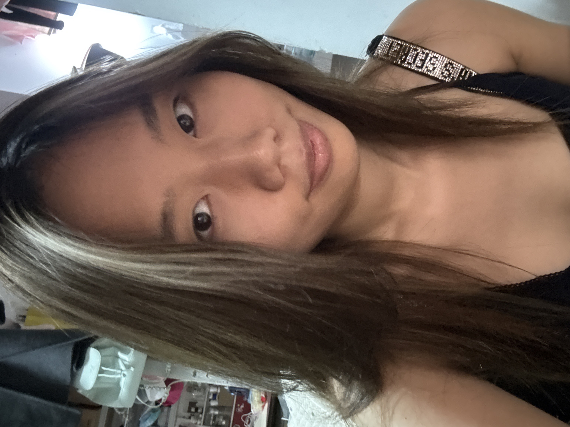 A woman with brown hair is facing the camera. She is wearing a black top with a rhinestone strap. The background appears to be a room interior, and there are blurred objects in the periphery. The location, Nan'an District, China, is not discernable from the image content.