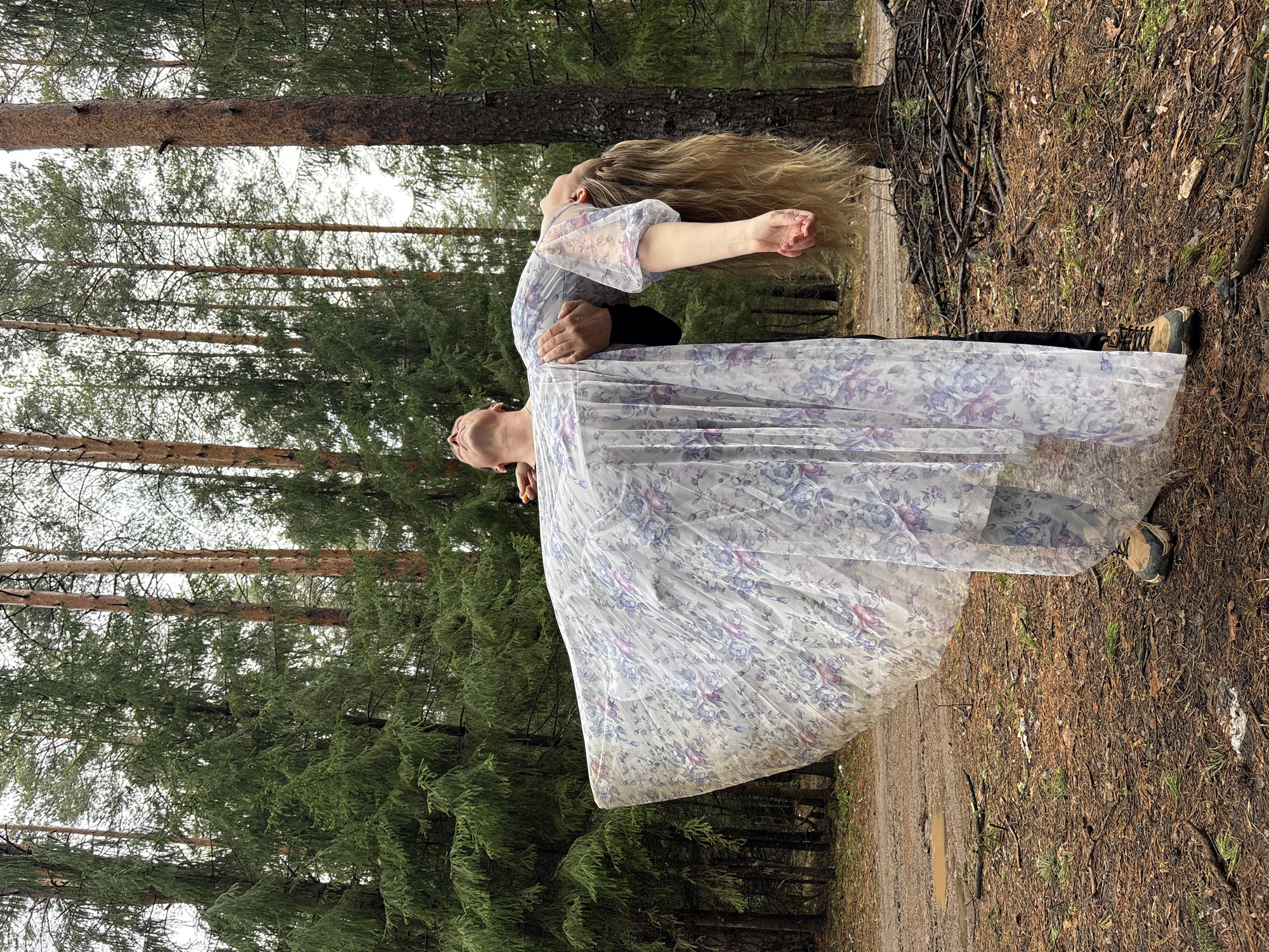 The image captures two individuals, a woman and a man, engaged in an artistic pose within a verdant forest in Chaikovsky, Russia. The woman, with long blonde hair flowing freely, is leaning backward with her head tilted skyward and arms outstretched, as if in mid-flight or a dance. She wears a translucent, flowing dress adorned with a delicate purple and blue floral pattern. The man, dressed in dark clothing and hiking-style shoes, supports her from behind, his hands around her waist.
The setting is a dense pine or coniferous forest, characterized by tall, straight tree trunks and a canopy of dark green needles. The forest floor is covered with a natural carpet of pine needles, leaves, and exposed earth, with a visible dirt path winding through the trees. The light suggests an overcast daytime, and the presence of small puddles on the path indicates recent dampness or rain. The overall scene evokes a sense of freedom, grace, and connection with nature. There is no visible text in the image.