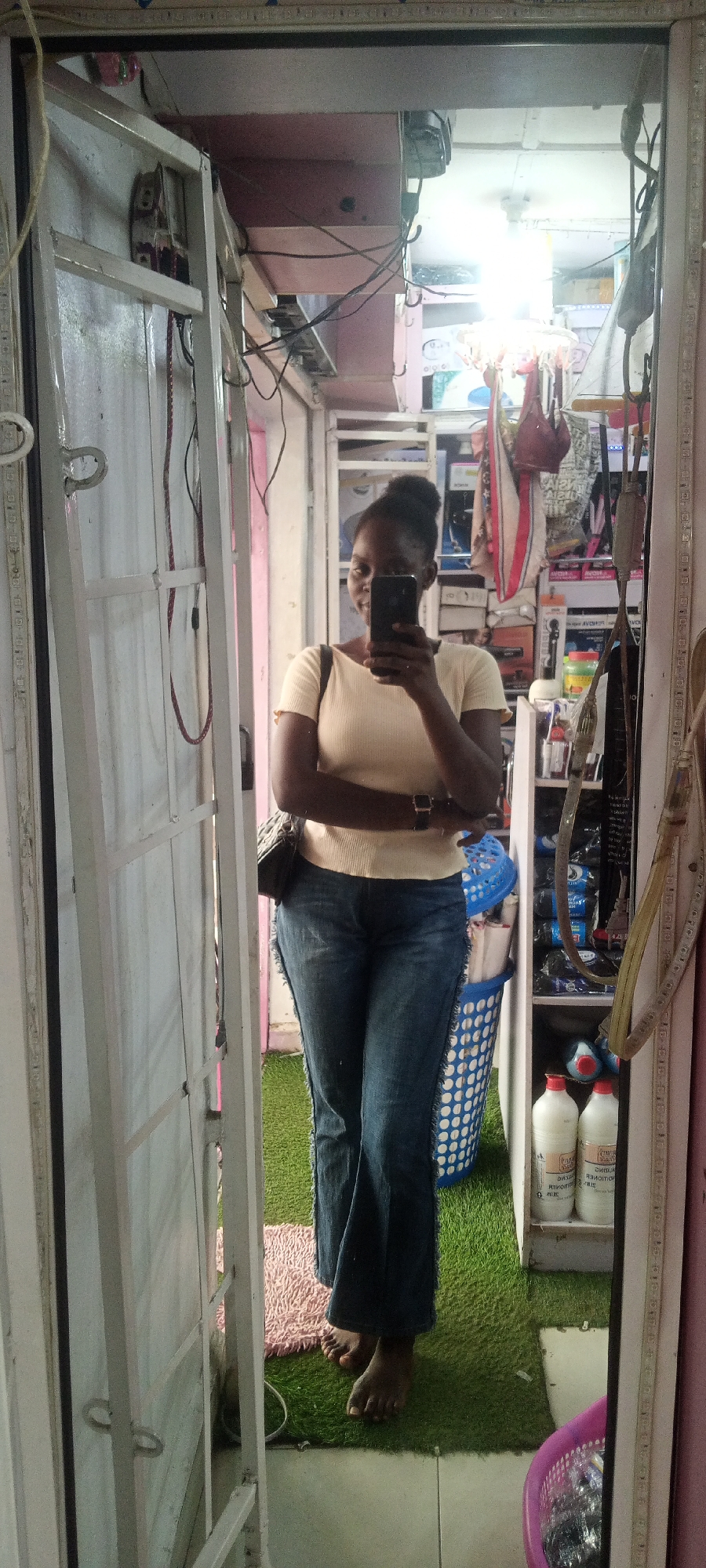 A young woman stands in front of a mirror, taking a selfie. She is wearing a light yellow, short-sleeved top and blue denim jeans. A black purse with a leopard print strap hangs from her left shoulder. She has her hair pulled up in a bun and is smiling faintly at the camera. She is barefoot and standing on artificial turf flooring.
The background appears to be a shop or a dressing room, with shelves filled with various items visible in the mirror's reflection. Among the items on the shelves are bottles of what appear to be hair products, hair dryers, and other personal care items. A blue laundry basket with white polka dots is visible behind her. The lighting suggests it is daytime, possibly indoors with artificial lights. The overall impression is casual and domestic, or perhaps a small retail space. The location is Jalingo, Nigeria.