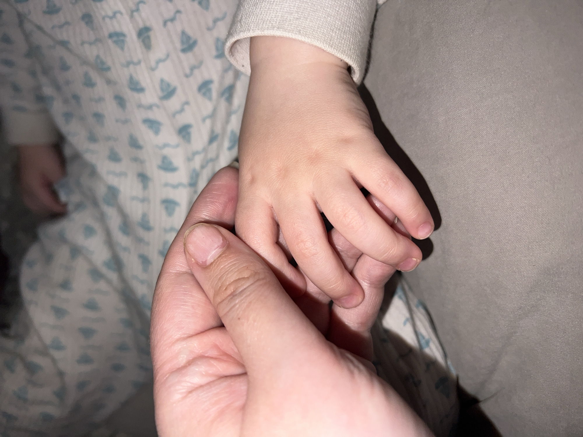 A close-up photograph shows an adult's hand holding a child's hand. The child is wearing a light-colored, long-sleeved garment, and part of a patterned blanket or sheet with a repeating boat design is visible. The adult's hand is open, cradling the child's hand, and is positioned against a neutral-colored surface, possibly clothing or bedding. The scene appears indoors. The city and country of Noviy, Russia cannot be determined from the information provided in the image.
