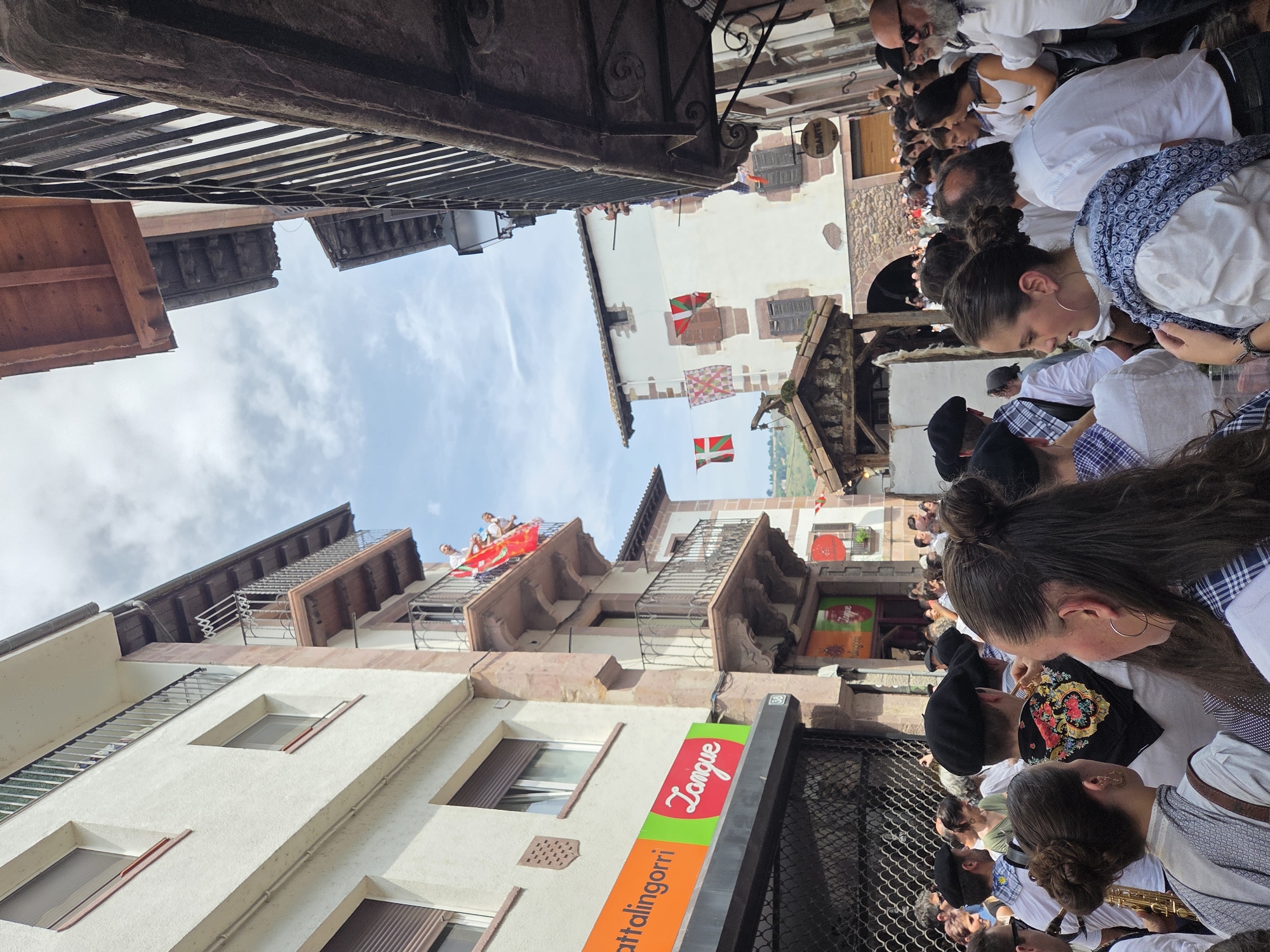 The image depicts a street scene in Baztan, Spain, with a crowd of people gathered outdoors. The architecture suggests a historic European town, with buildings featuring balconies, stone accents, and wooden eaves. Several flags, including the Basque flag, are visible, indicating a cultural event or celebration. People are dressed casually, with some wearing traditional Basque berets. The atmosphere appears lively, with a sense of community and festivity. A street-level view shows shops with signage, and the overall scene is bathed in natural daylight, suggesting it was taken during the day.