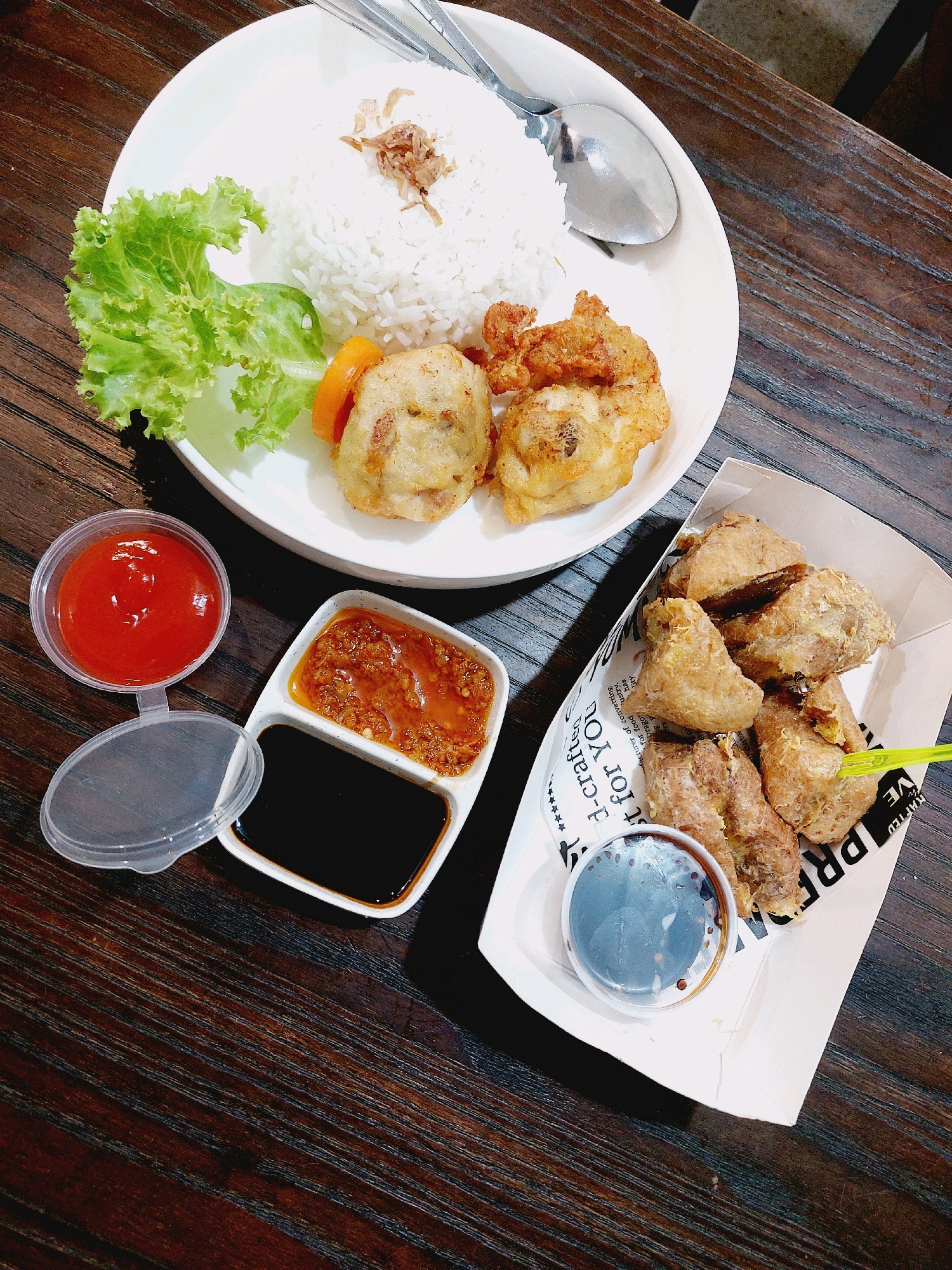 The image shows a close-up, overhead view of a meal served on a dark wooden table. The meal consists of white rice, fried food items, and several dipping sauces. On a white plate, a mound of white rice is topped with fried shallots. To the left of the rice is a piece of lettuce and some sliced orange items, possibly carrots. Beside the rice are two fried patties. To the left of the plate, there's a small, clear plastic container with a lid, filled with a red sauce, likely ketchup. Next to this is a divided dish with two compartments: one contains a reddish-brown sauce, possibly chili paste, and the other contains a dark liquid, likely soy sauce. To the right of the plate, in a white paper tray lined with text, are several pieces of golden-brown fried food, appearing to be tofu or some other battered and fried ingredient. A small, clear plastic container filled with a dark liquid, possibly a dipping sauce, sits in the paper tray. A bright green plastic utensil is also visible in the tray. The lighting suggests an indoor setting. The food is presented in a casual, possibly restaurant or street food style. The environment appears to be the city of Tarakan, Indonesia, given the context of a common meal presentation. There are no people visible in the image.