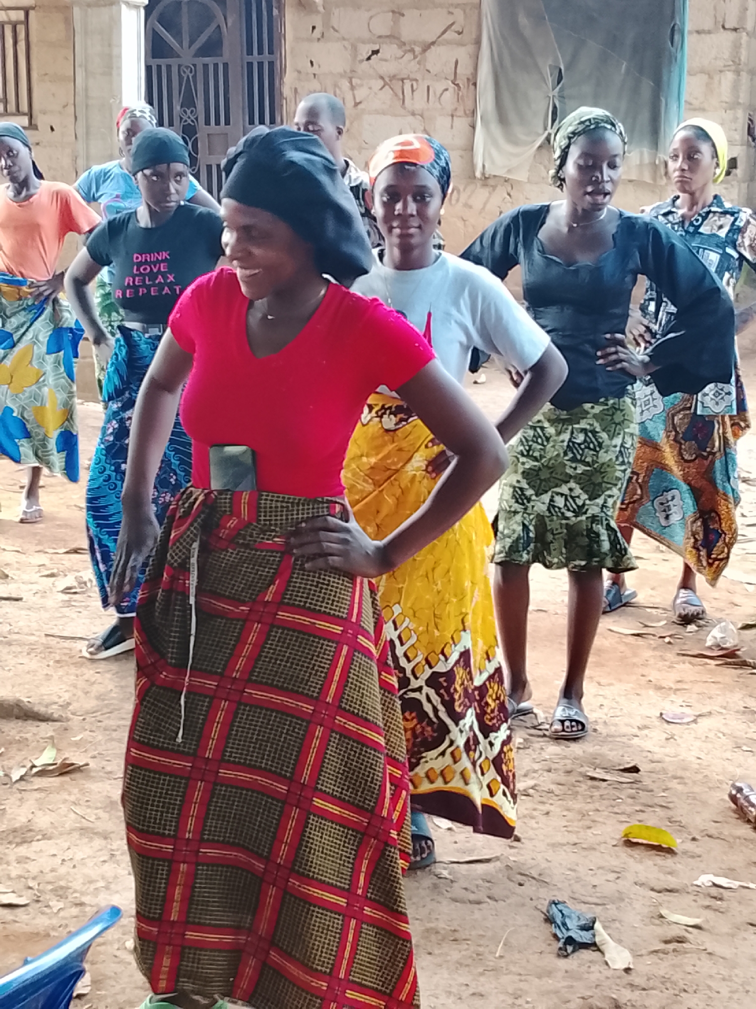 In Akwanga, Nigeria, a group of women are captured in a vibrant outdoor scene, likely engaged in a lively activity. The setting appears to be a dusty, open area, possibly a courtyard or village space, with a building in the background featuring a rough-hewn wall and a metal-barred doorway. The ground is dirt, scattered with a few dry leaves and some discarded items, suggesting a casual, everyday environment.
The central focus is on a group of women, many of whom are wearing colorful wrappers and headscarves. Their stances and expressions suggest movement and engagement. The woman in the foreground, wearing a red top and a plaid skirt, smiles broadly and looks to her left, her hand on her hip. To her left, another woman in a black t-shirt with white text, also with her hand on her hip, looks forward with a determined expression. She is wearing a black headscarf. Behind her, a woman in a yellow wrapper and a white top looks towards the camera with a neutral expression, also wearing a patterned headscarf. Further back, other women are visible, some with their hands on their hips, their attire contributing to the lively, communal atmosphere.
The clothing of the women is a notable detail, with various patterns and colors of wrappers and headscarves creating a visually rich tapestry. One woman's black t-shirt has visible white text that reads "DRINK LOVE RELAX REPEAT". The time of day is suggested by the natural lighting, which appears to be bright, implying daytime, possibly late morning or early afternoon. The overall mood conveyed is one of energy and community.