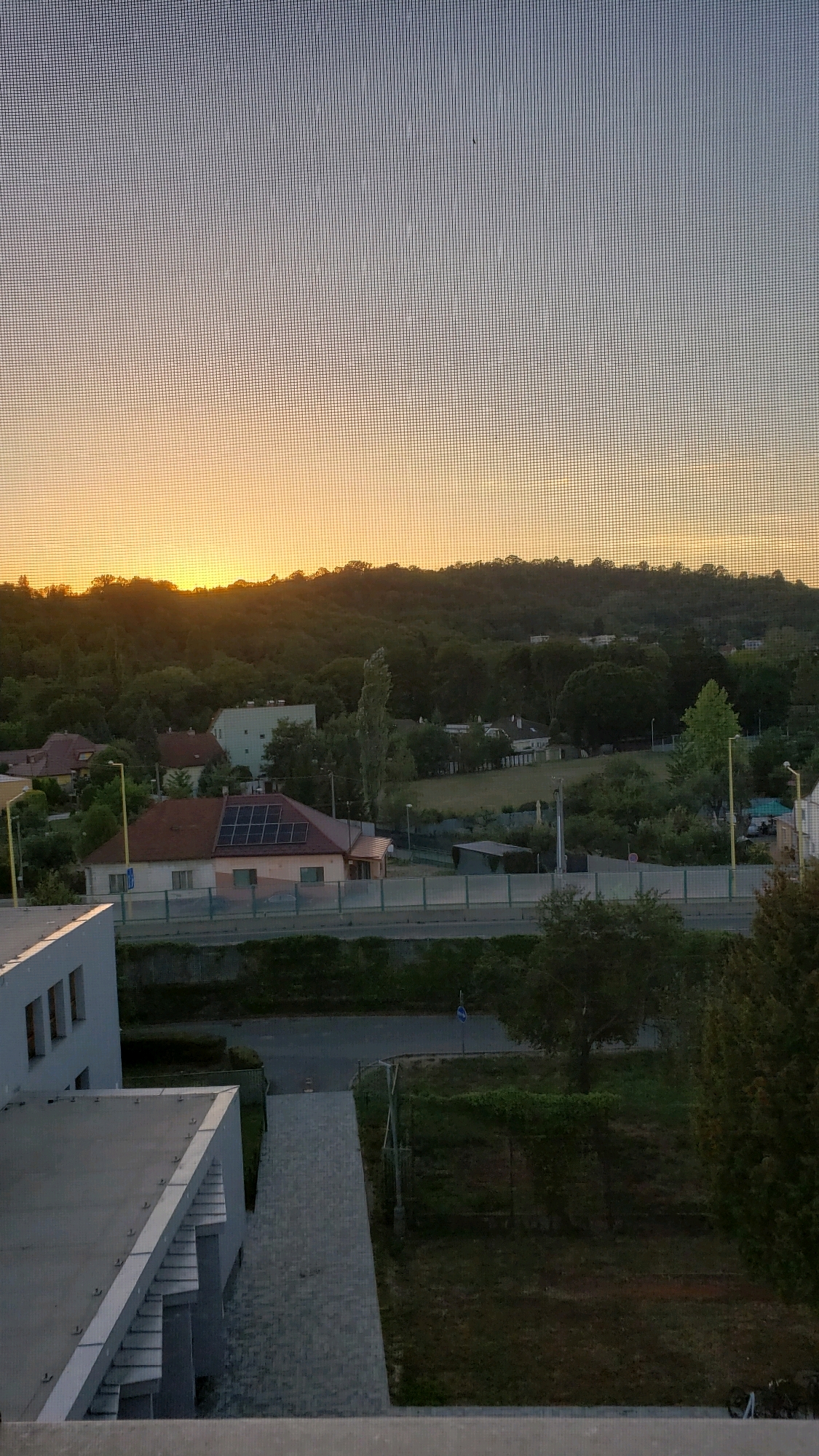 The image shows a view through a window screen of a residential area in Prešov, Slovakia. The sky is a gradient of soft orange and blue, indicating either sunrise or sunset. A treeline with a rounded hill forms the horizon. Below the hill, there are several houses, some with red tile roofs. One house in the foreground has solar panels installed on its roof. A road runs horizontally across the middle of the frame, with a fence separating it from a grassy area. In the foreground, there is a modern building with a grey roof and white exterior. The overall scene depicts a peaceful, suburban landscape.