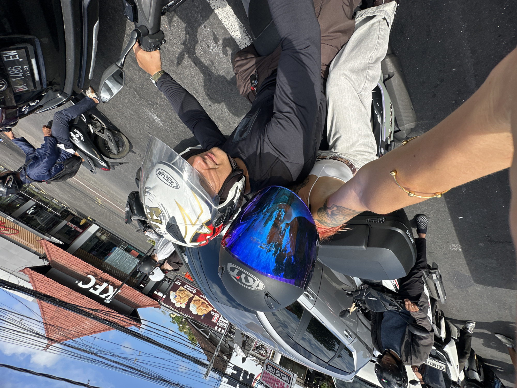 Here is a detailed description of the image:
**Scene Overview:** The image appears to be taken in Kuta Selatan, Indonesia, judging by the license plate details ("DK" for Bali). The photo is a street-level shot, possibly a selfie, capturing a scene with people on motorbikes, cars, and a KFC restaurant in the background.
**People:**
* There is at least one person who is clearly visible and posing for the photo. They are wearing a helmet and appear to be sitting on a motorcycle.
* A part of an arm with a tattoo and a gold bracelet is visible, likely belonging to the person taking the picture.
* Other individuals can be seen in the background, some on motorbikes and others near the KFC.
**Objects:**
* Several motorbikes are present.
* Helmets are visible; one person is wearing one, and others are placed on a vehicle.
* A car or van is visible near the person taking the photo.
* A KFC restaurant is in the background, with visible signage.
**Actions/Interactions:**
* The central person is posing for a picture.
* Other individuals are moving around in the background, likely going about their daily activities.
**Environment:**
* The setting is an urban street with buildings and traffic.
* The weather appears to be sunny, casting shadows.
* There are overhead power lines visible.
* The presence of a KFC and the license plates suggest the location is Kuta Selatan, Indonesia.
**Specific details:**
* The license plate on one of the motorbikes has the letters "DK" indicating it's registered in Bali.
* A KFC restaurant is identifiable in the background.
* At least one of the helmets is a KYT brand.
