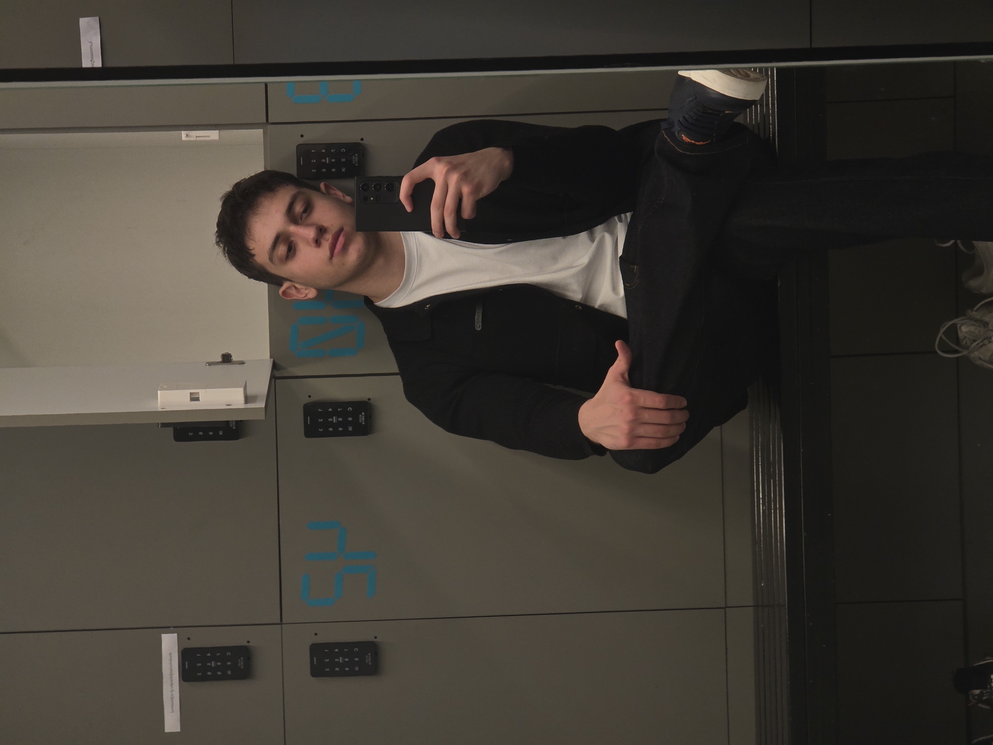 A person is taking a selfie in a mirror in front of lockers, possibly in Yerevan, Armenia. The lockers are gray with blue numbering and digital keypads. The person is wearing a white t-shirt, a black jacket, and dark pants with sneakers. The mirror reflects the person's image and the surrounding locker area.