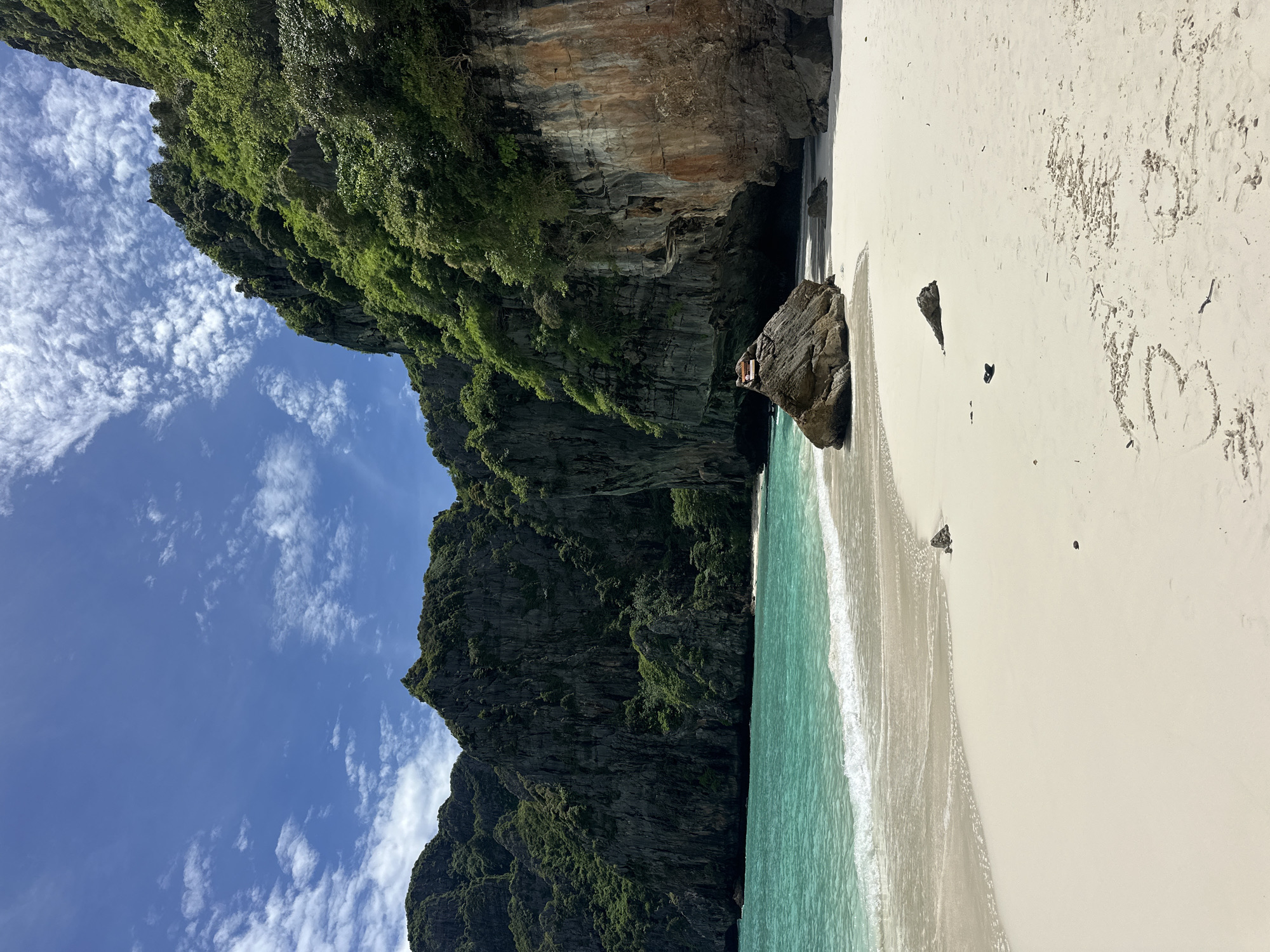 A stunning view of a pristine white sand beach meets the turquoise waters of the Andaman Sea. Towering limestone cliffs, lush with green vegetation, rise dramatically from the water's edge, forming a breathtaking backdrop. The sky above is a vibrant blue, dotted with scattered white clouds. On the shore, a large boulder sits prominently in the sand, with a small dark object resting on top. Further back, a cave is visible within the cliff face. The waves gently lap at the shore, leaving a frothy white edge on the sand. The overall scene evokes a sense of tropical paradise and natural beauty, likely a secluded cove or bay accessible from Ao Nang, Thailand.