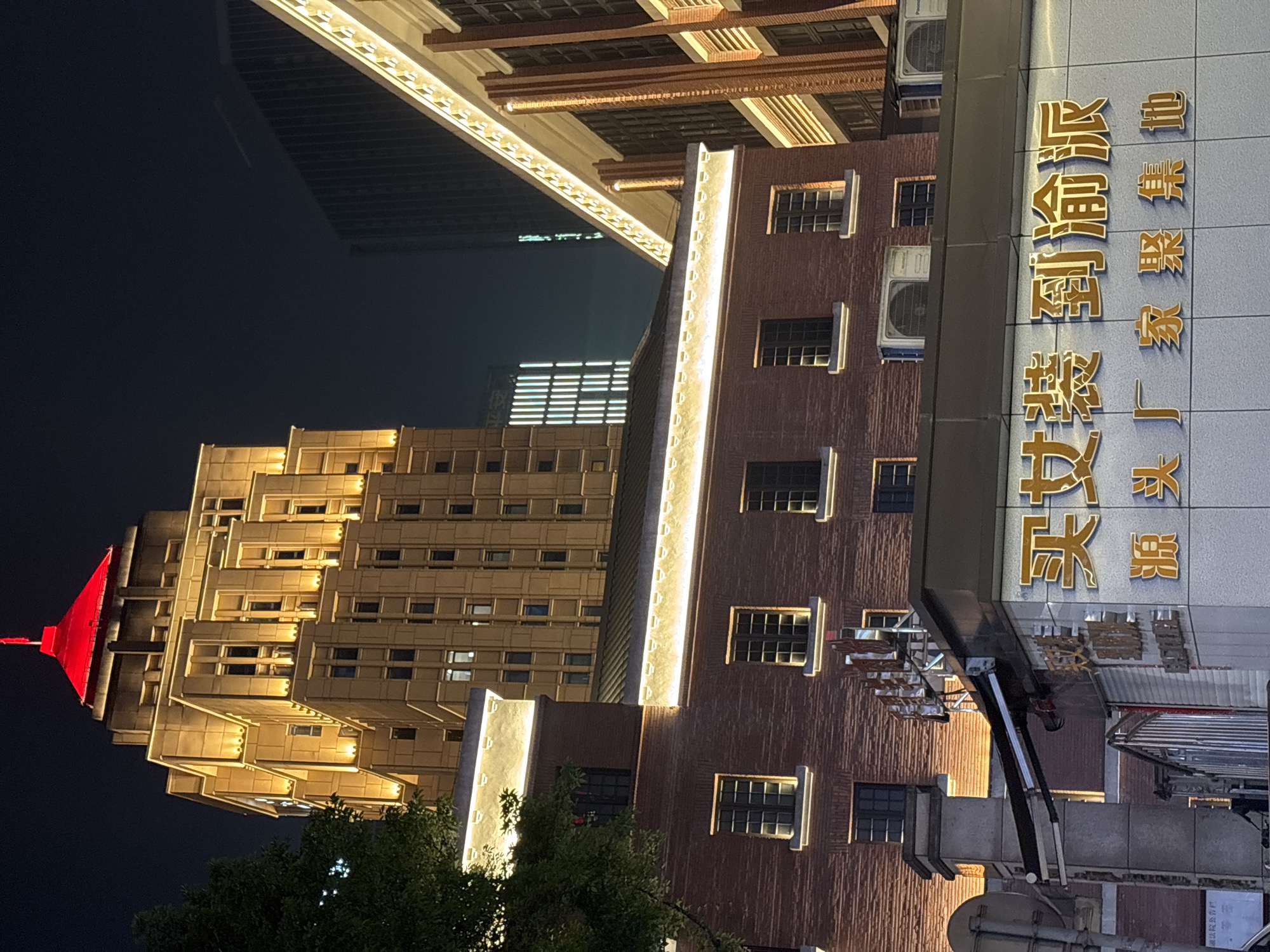 The image depicts a nighttime scene in the Yuzhong District of Chongqing, China. A tall, illuminated building with a red spire is visible on the left. Adjacent to it is a brick building with multiple windows. A sign with Chinese characters, illuminated in gold, is attached to the right side of the brick building. Overhead is a structure with lights. The environment is urban.