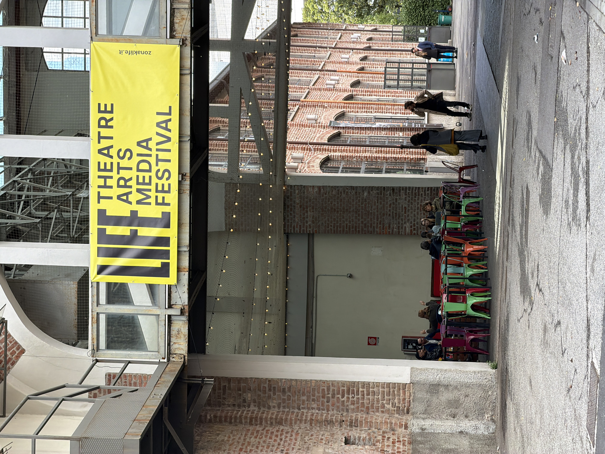The image captures an outdoor scene in Unknown, Unknown, featuring architectural elements, people, and a festival banner. A large yellow banner hangs on the left, displaying the words "THEATRE ARTS MEDIA FESTIVAL" vertically in black text. The banner's website is zonaklife.it. A complex structure of metal beams and brickwork surrounds the banner. In the background, a multi-story brick building with arched windows and decorative brickwork is visible. Below this structure, several people are seated at colorful plastic chairs and tables, engaging in conversations and activities. Further along the pathway, additional people are walking or standing. The environment appears to be a passageway or alleyway, possibly part of a festival location or event space. The overall lighting is subdued, suggesting an outdoor setting with partial cover or shade.