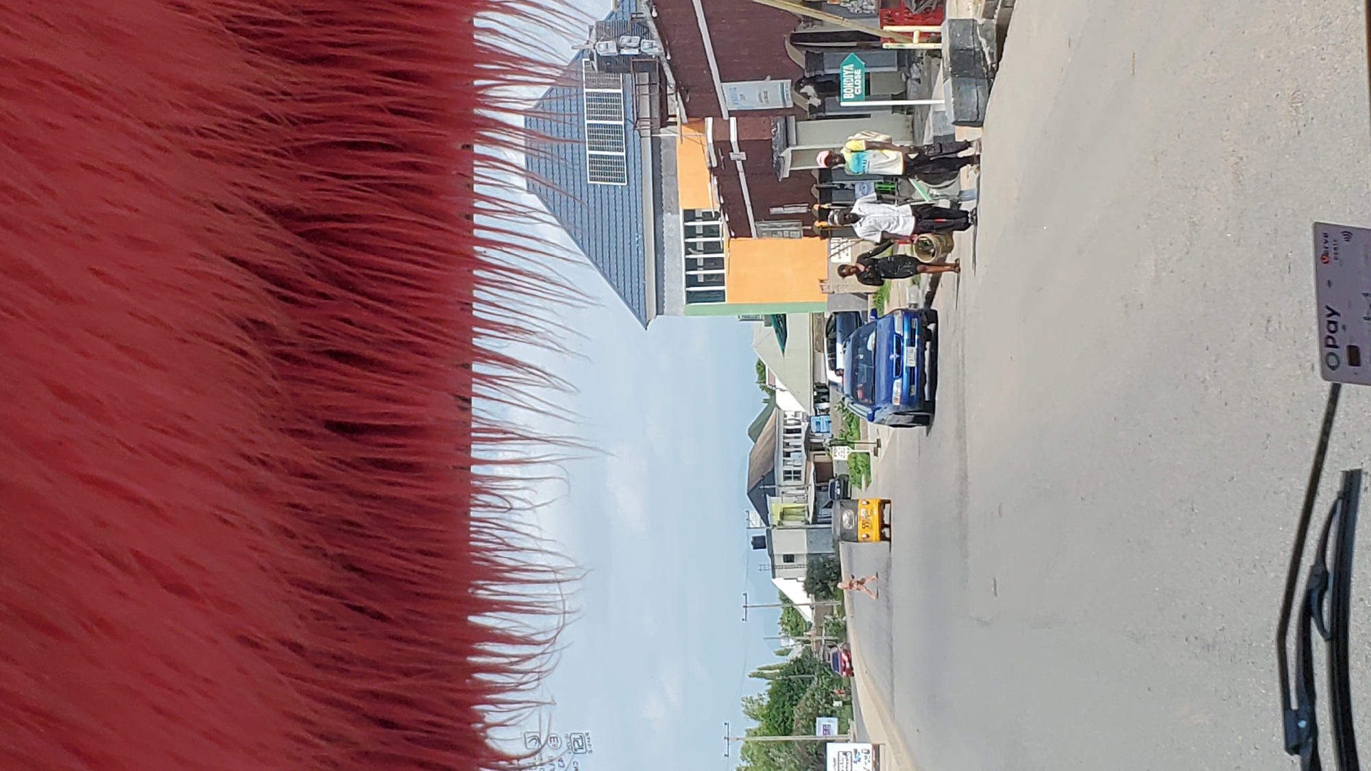The image captures a street scene in Jalingo, Nigeria, viewed from inside a vehicle. The foreground is dominated by a furry, pink object, likely a car interior accessory, hanging from the top. The road ahead shows a slight curve and is paved with asphalt. Several vehicles are present: a yellow auto-rickshaw, a blue sedan, and a few other cars further down the road. Pedestrians are also visible, including two men and one woman walking on the side of the road. Buildings line the street, with one prominently featuring an orange and green facade and a blue roof. A green directional sign that reads "BONDIYA CLOSE" is also visible. The sky is cloudy, suggesting an overcast day. The overall impression is of a typical urban street in Nigeria.