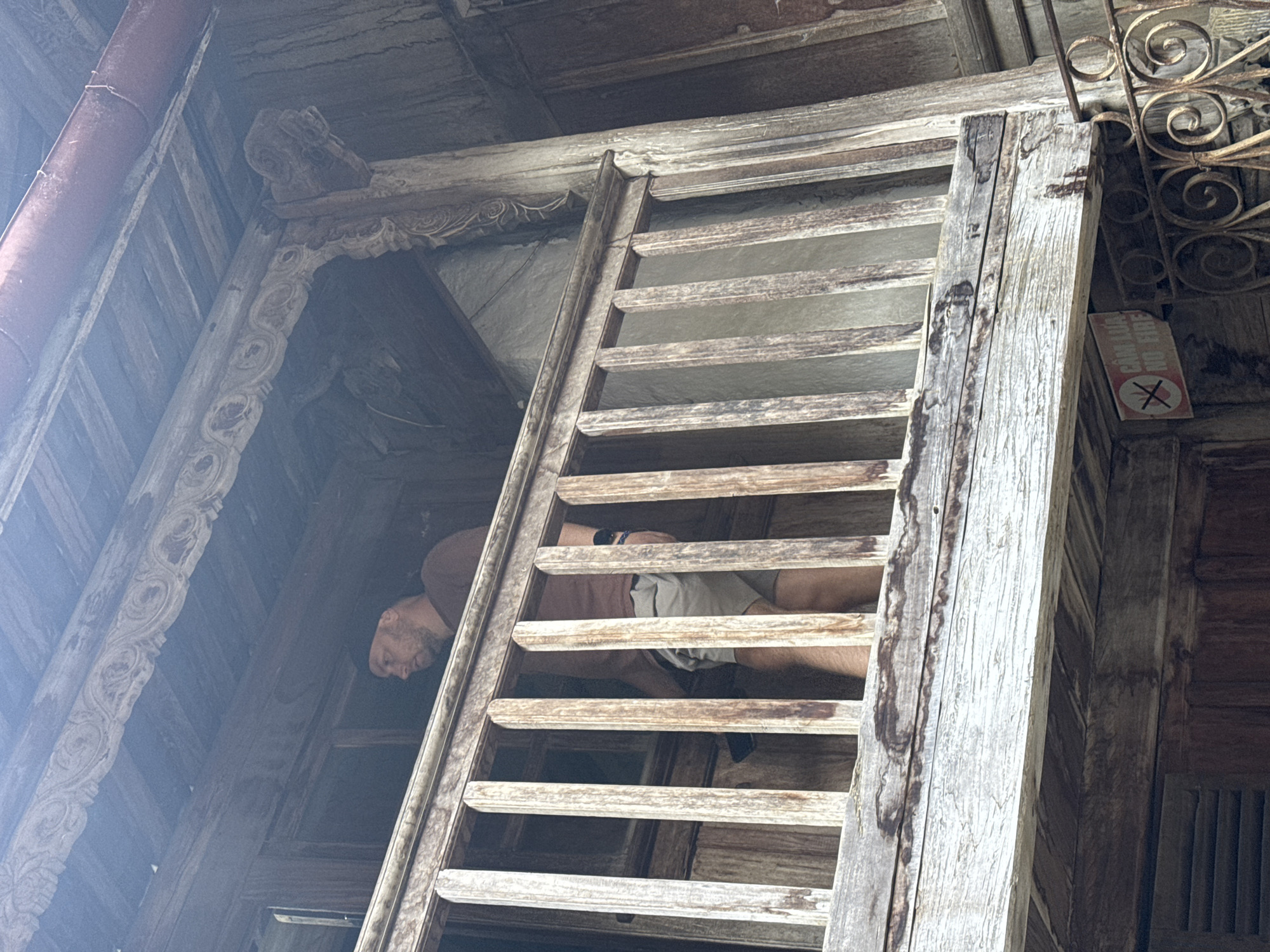A low-angle shot reveals a weathered wooden structure, possibly part of a building, in Sà Phìn Commune, Vietnam. A man is visible behind a ladder-like series of horizontal wooden slats; his head is visible on the left side of the slats and his torso is obscured in the slats. A sign with a no smoking symbol is affixed to the wall to the right of the structure. The surroundings appear to be made of aged wood with intricate carvings.