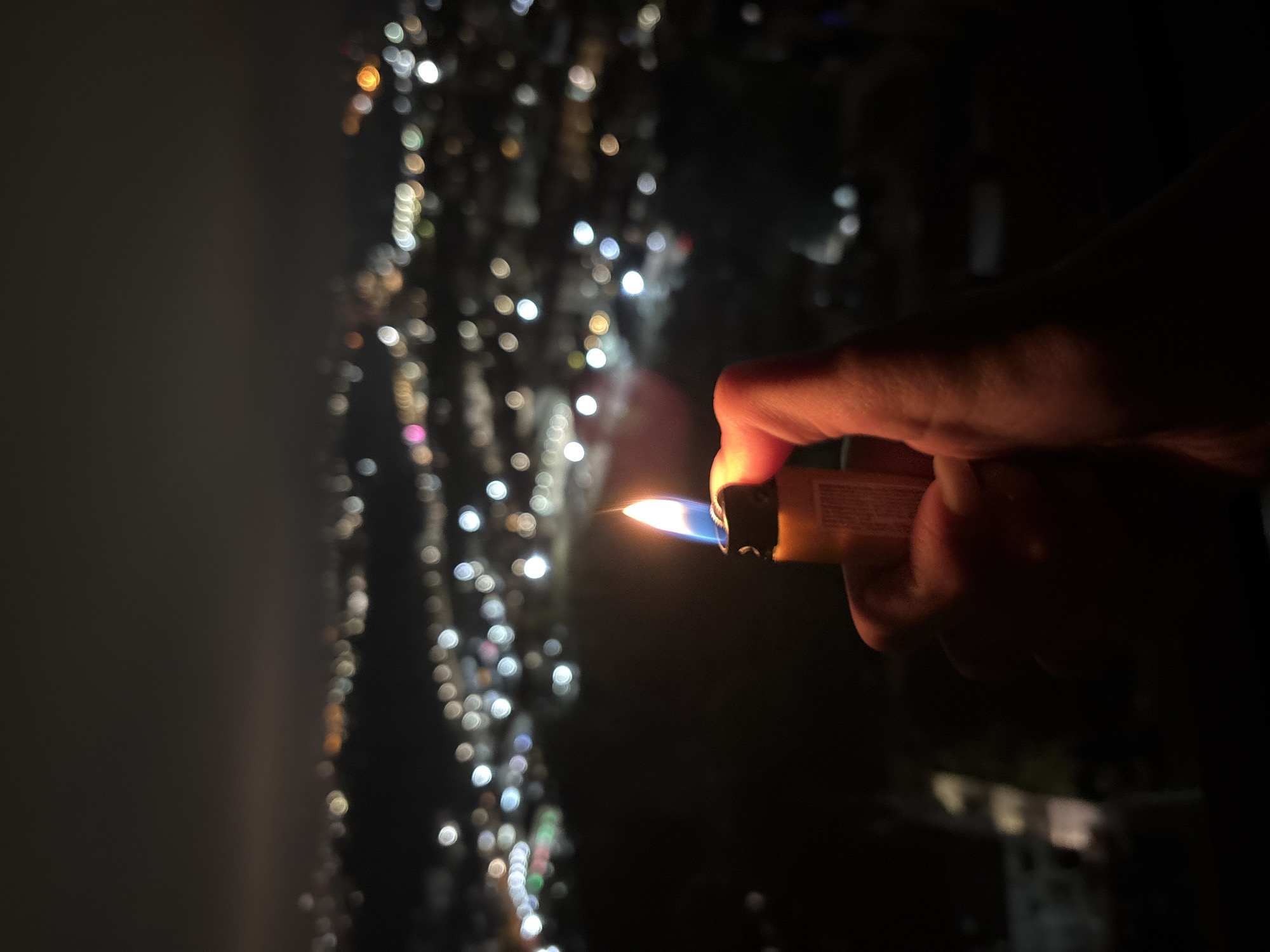 A close-up, low-angle shot shows a person's hand holding a lit lighter against a blurred backdrop of city lights. The hand, appearing somewhat shadowy, holds a yellow disposable lighter with a visible blue and yellow flame. The background consists of blurred city lights, suggesting an urban environment at night, specifically the city of Sochi, Russia. The lights appear as bokeh circles, varying in color and brightness. The overall mood is dark and slightly mysterious, highlighting the small flame against the distant city illumination.